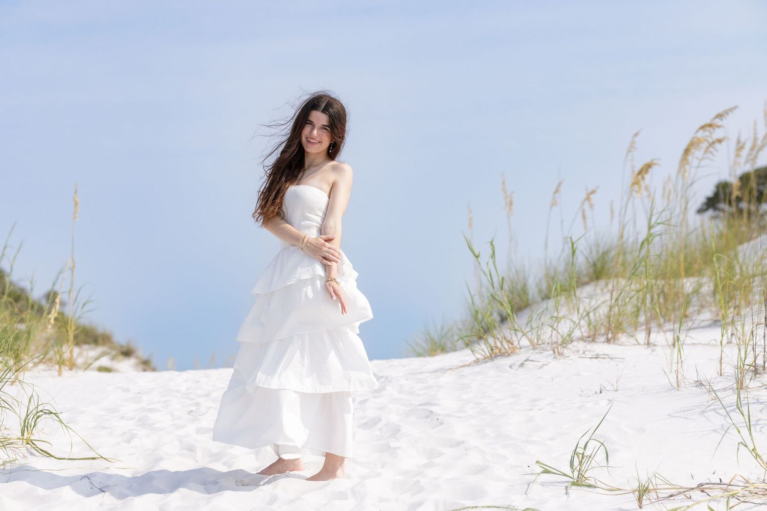 Young woman in white strapless tiered dress posing barefoot on sandy beach dunes with sea oats.