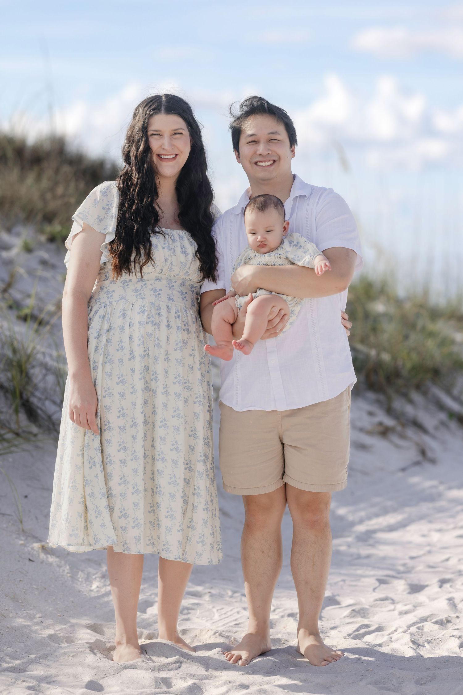 Happy couple holding their newborn baby on a sandy beach with dunes in background.