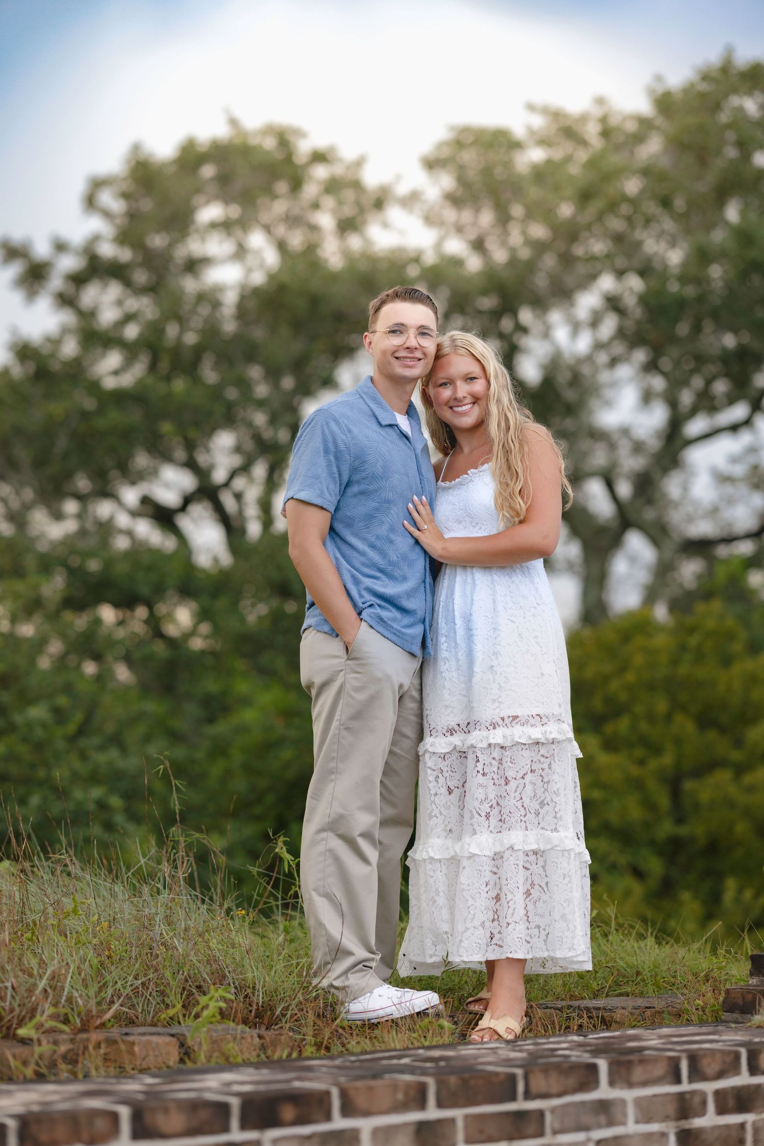 A smiling couple poses outdoors, man in blue shirt and khakis, woman in white lace dress, with green trees behind them.