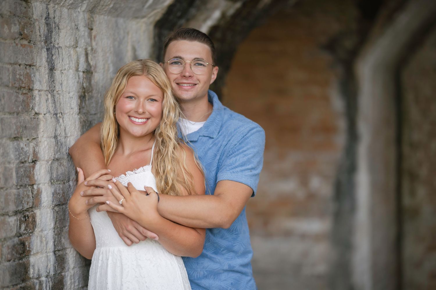A smiling young couple embraces against a rustic brick wall, the woman in white and man in blue shirt.
