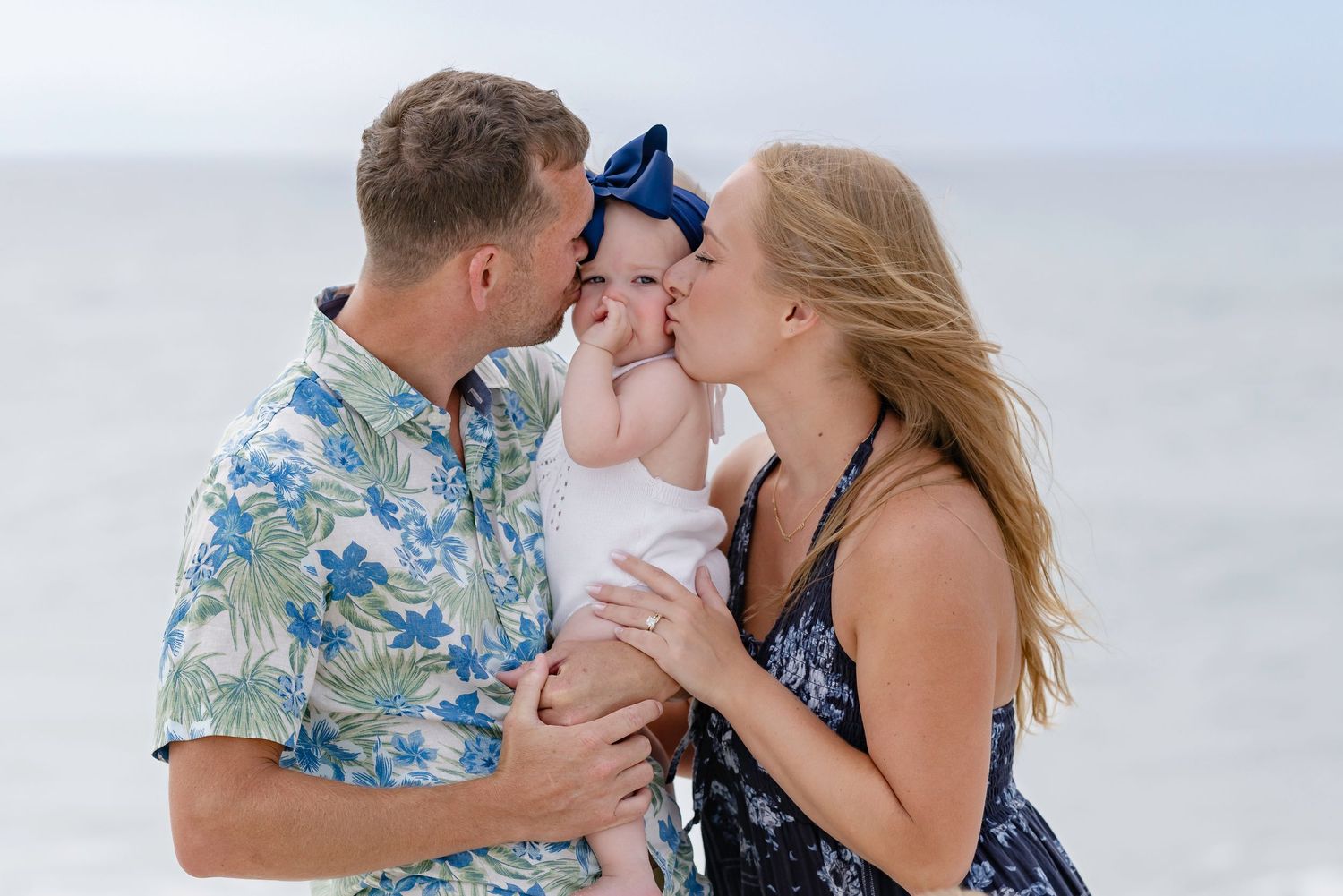 A couple kisses their baby girl on the cheeks at the beach, dad in floral shirt, mom in navy dress.