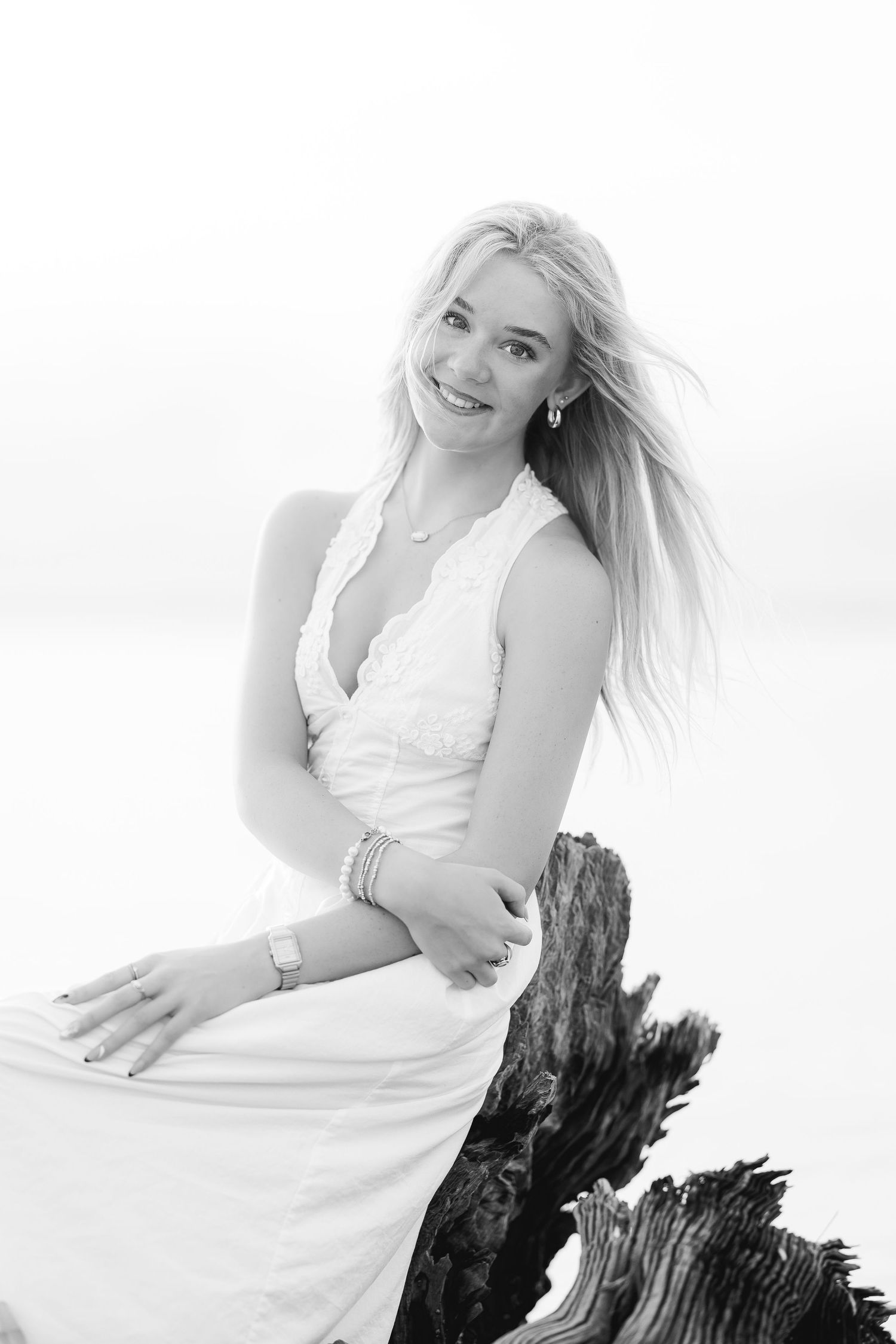 Smiling blonde woman in white dress sitting on driftwood at beach, black and white portrait photography.