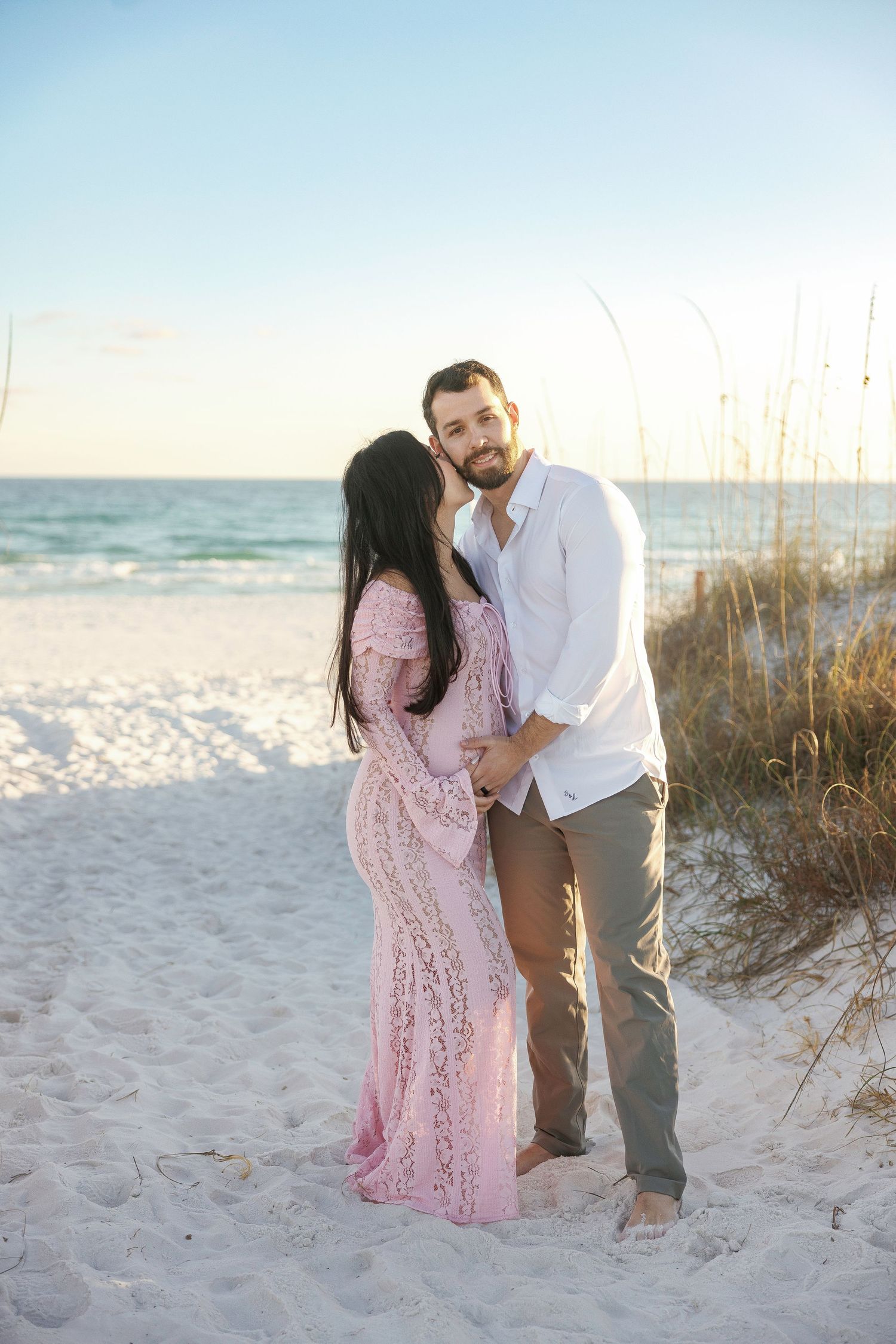 A couple shares a romantic moment on a white sandy beach with sea oats and ocean waves in the background.