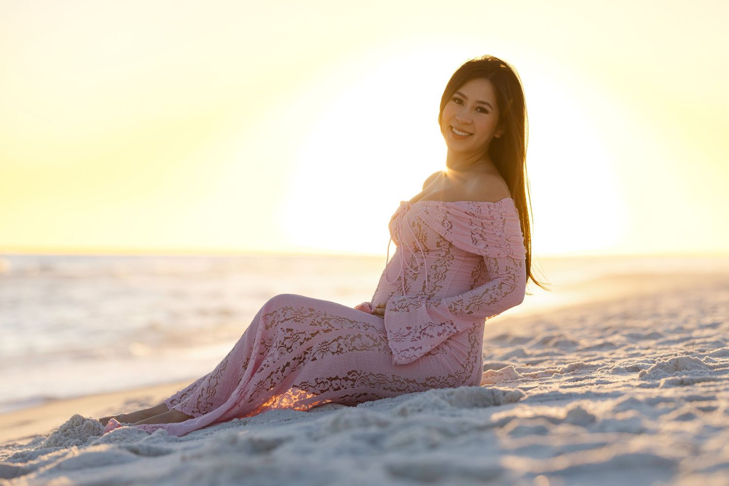 A person in a flowy pink dress sits gracefully on a sandy beach at sunset, creating a dreamy coastal scene.