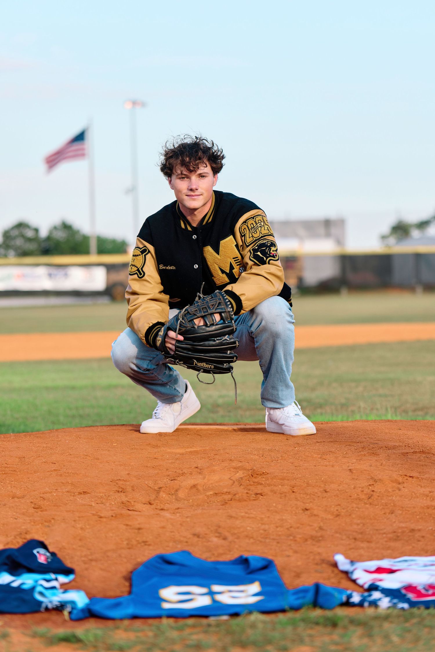 Baseball player crouches on infield dirt with blue jerseys laid out in front during sports photography session.
