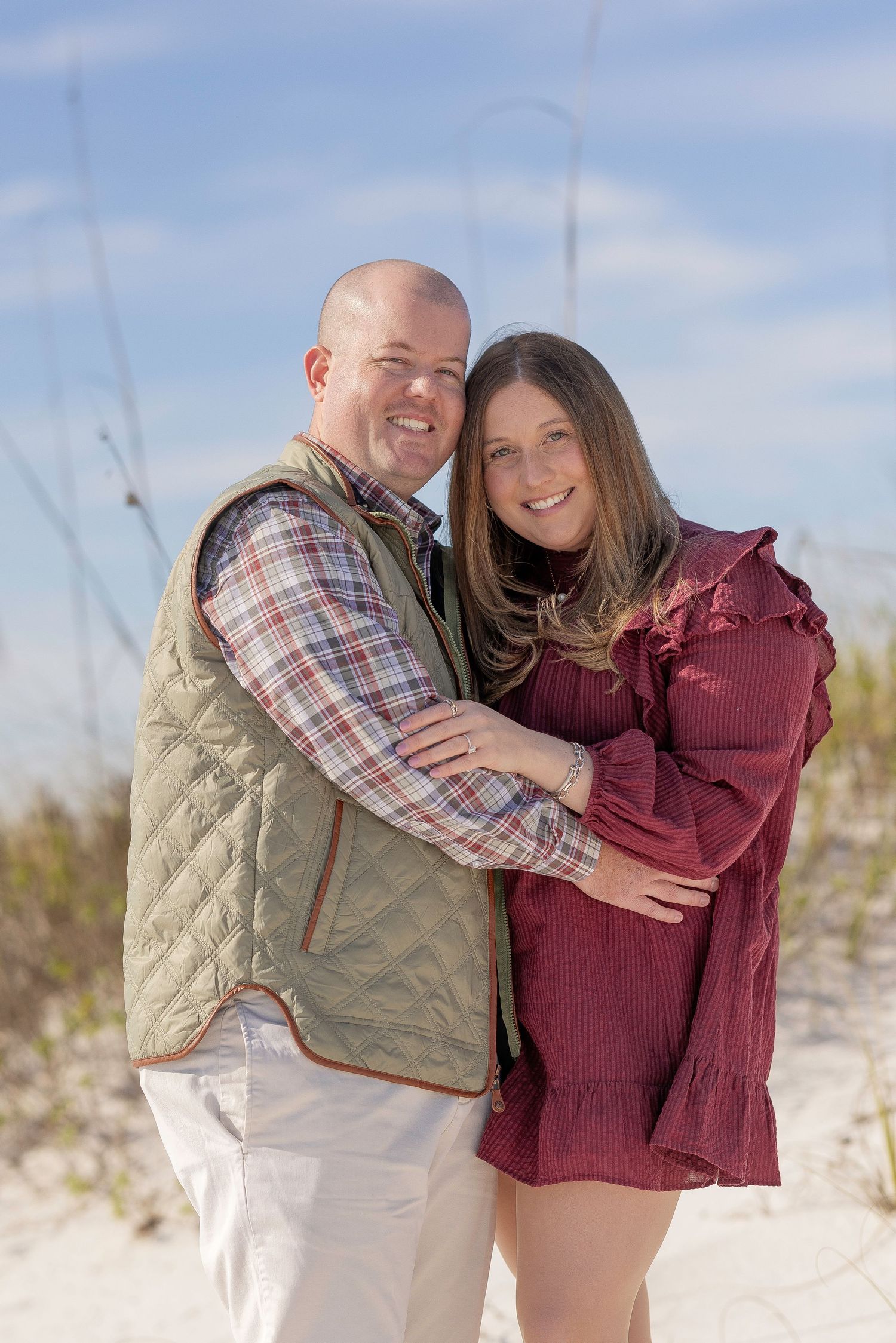 A couple embraces on a white sandy beach wearing casual fall attire against a bright blue sky.