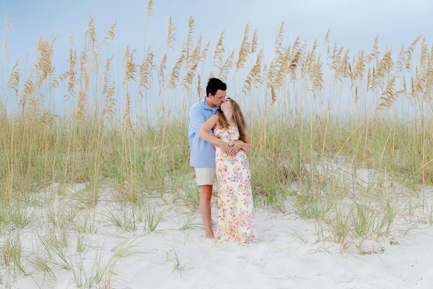 A couple embraces while standing among tall beach grass and sand dunes on a serene summer day.