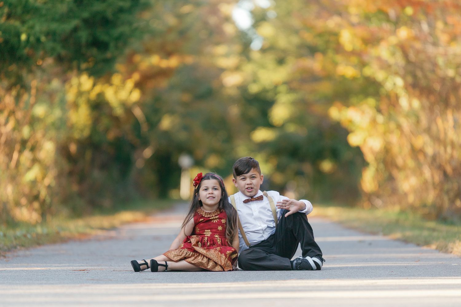 Two children lay on a rural road surrounded by autumn trees and golden sunlight.