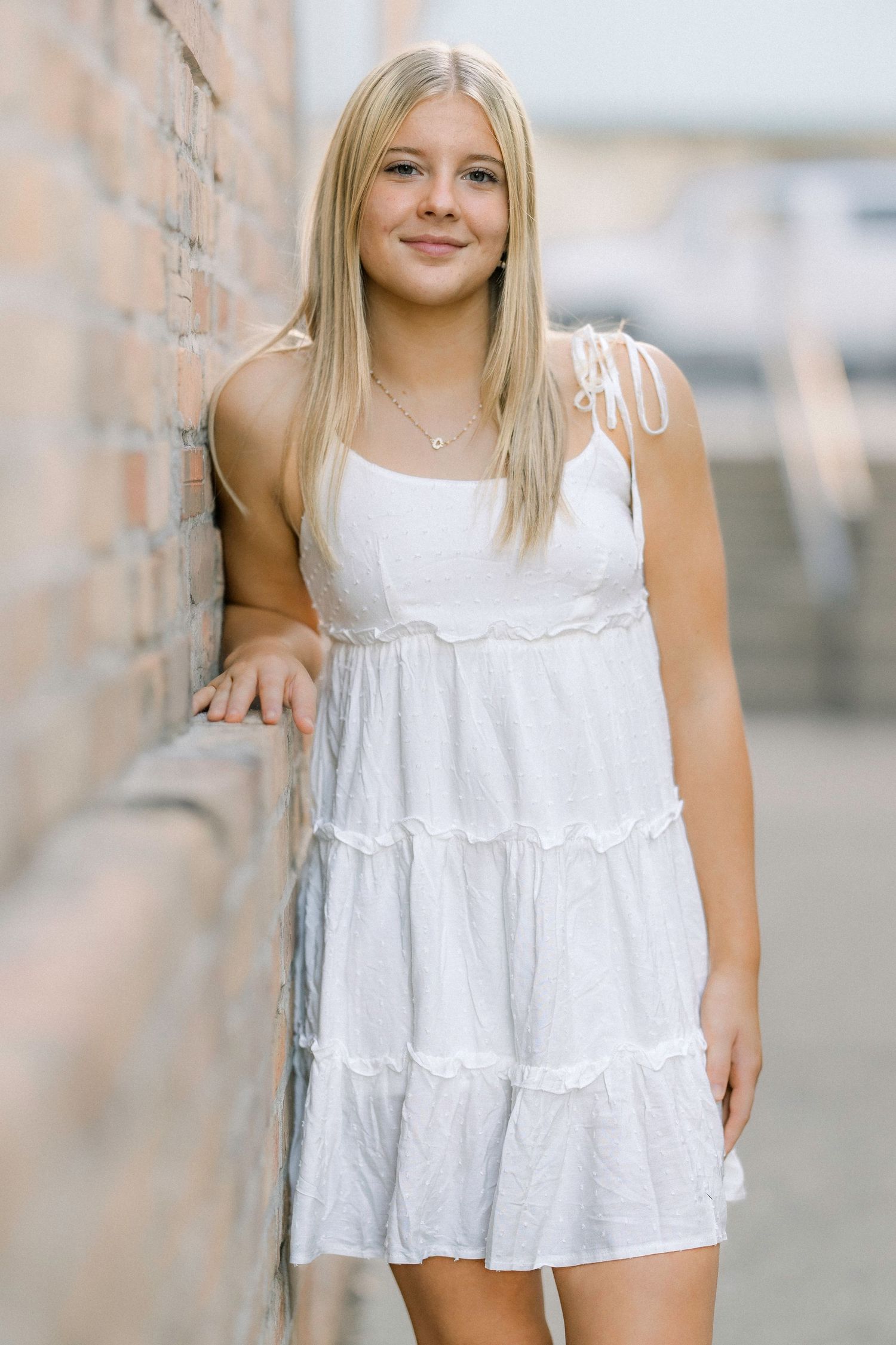 Young blonde girl in white tiered sundress leaning against brick wall, smiling outdoors.