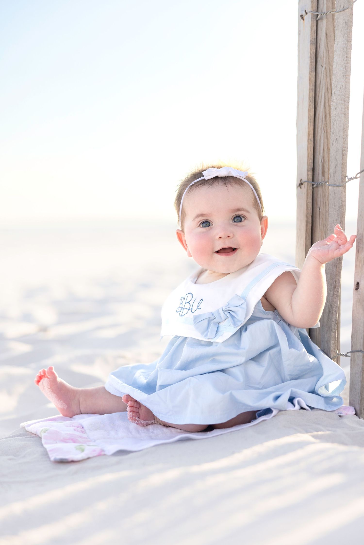 Adorable baby girl in blue dress and headband sitting on white sand beach near wooden fence.