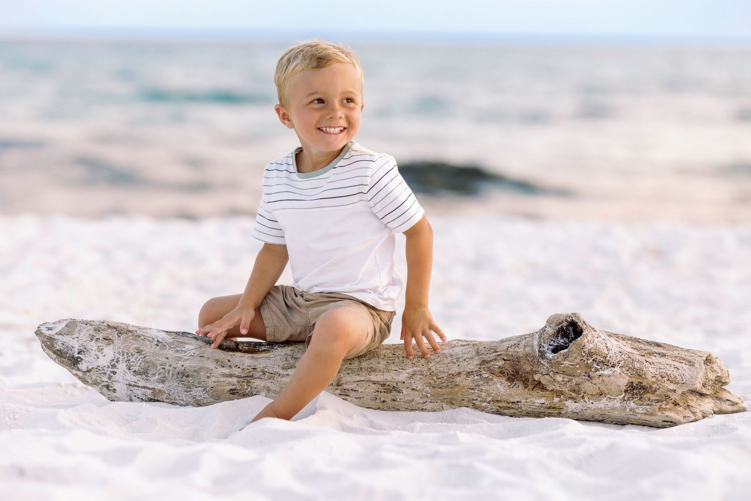 Happy young boy sitting on driftwood on a white sand beach, smiling in a striped shirt and shorts.