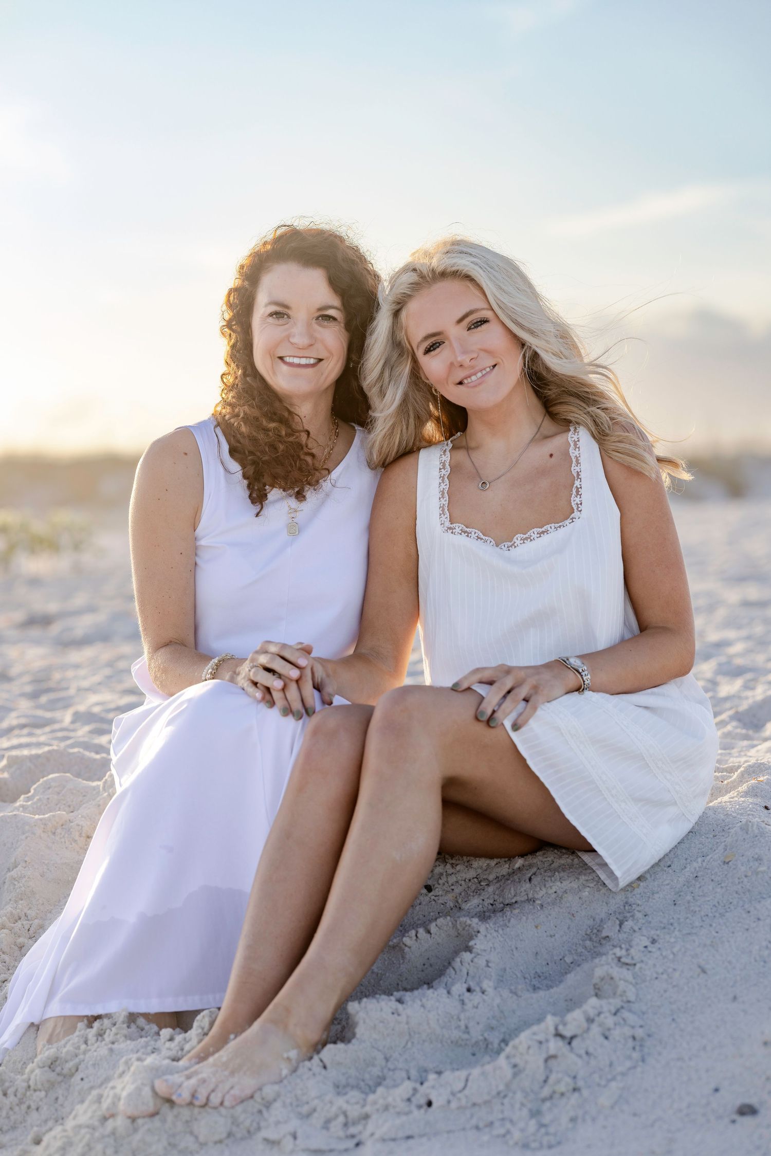 Two smiling women in white dresses sit together on sandy beach at golden hour sunset.