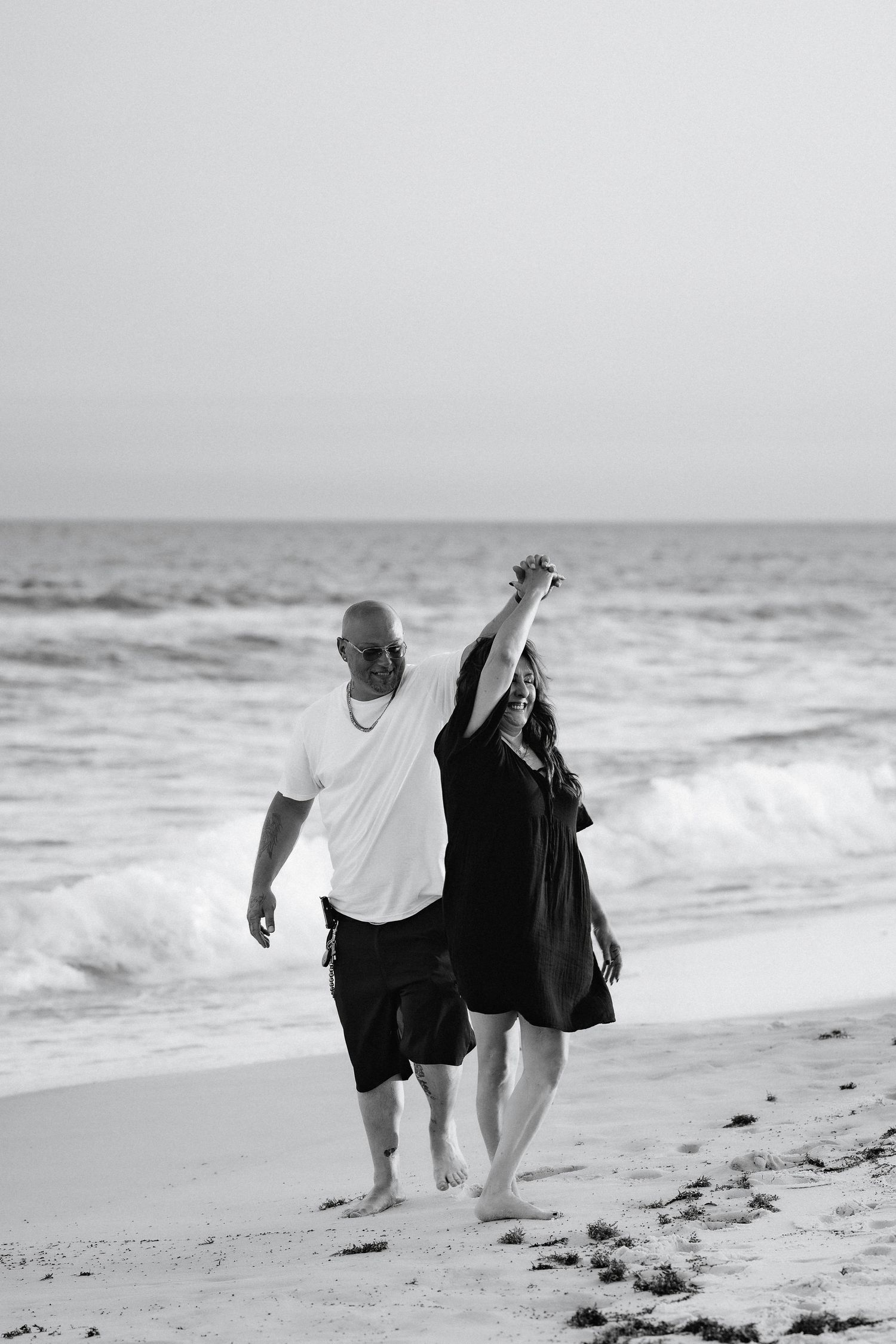 A black and white photo of a couple holding hands while walking along a beach with ocean waves.