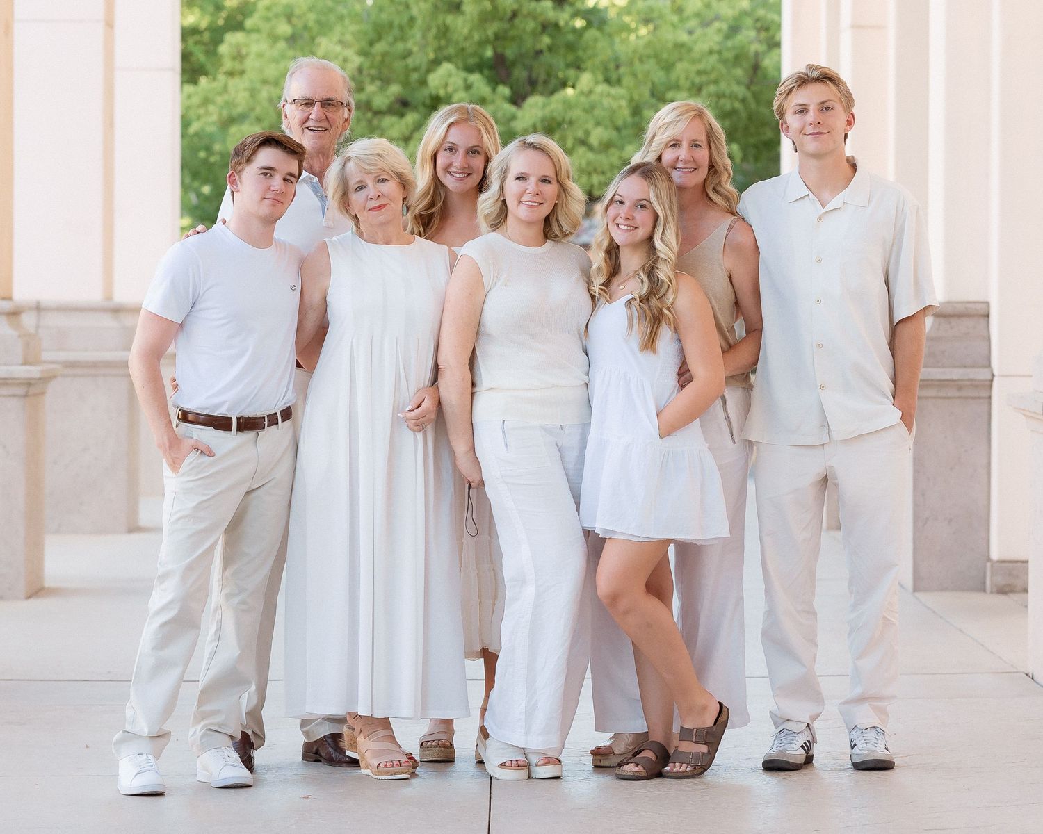 A cheerful family of eight poses together in coordinated white and beige outfits outdoors near elegant columns.