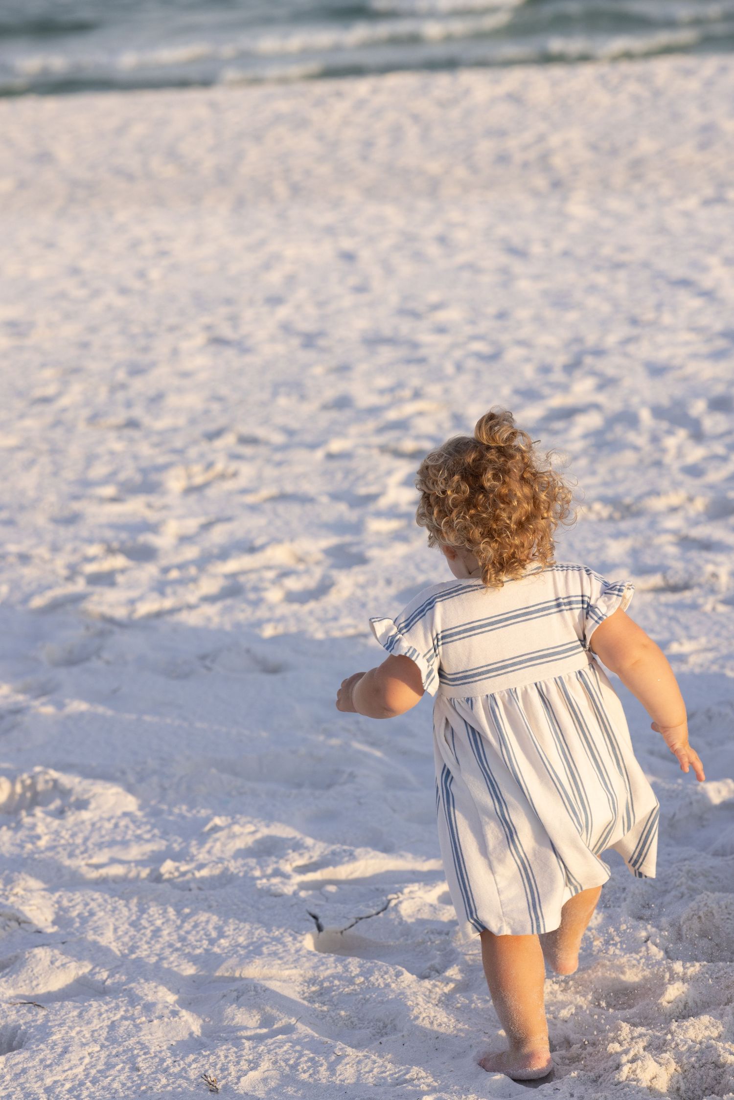 A toddler girl in a striped dress runs toward the ocean on a white sandy beach at golden hour.