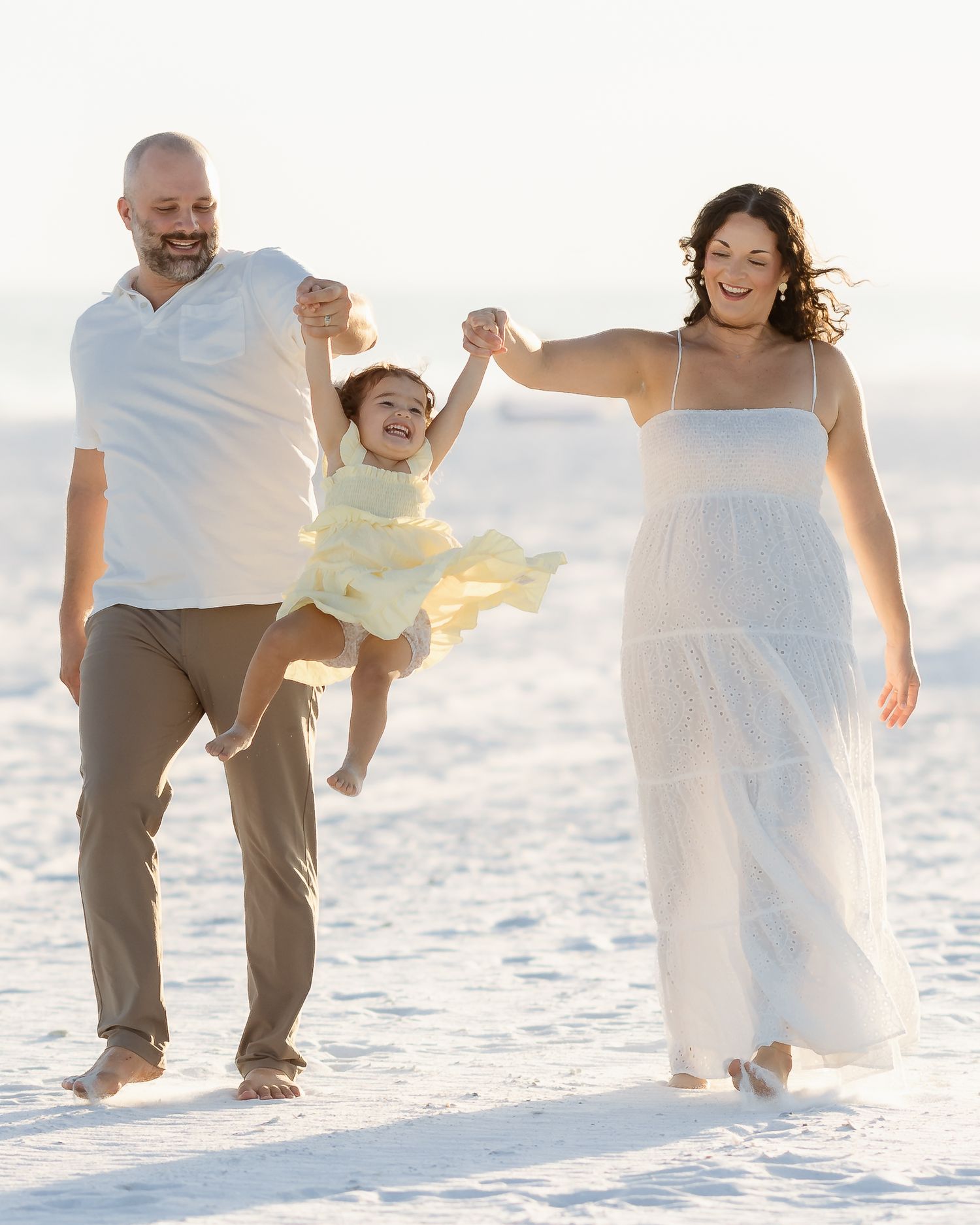 Family of three walking on a sunny beach, parents swinging their laughing child between them, wearing white and yellow outfits.