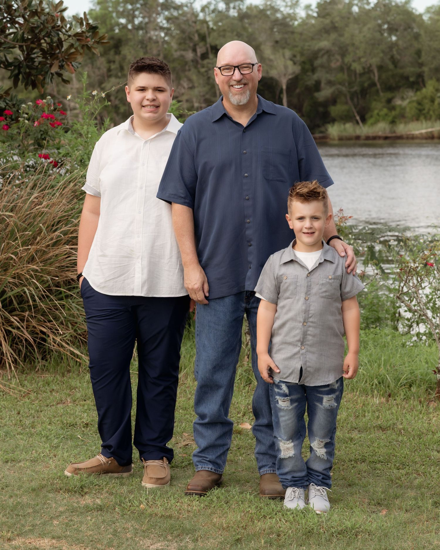 A bald man in a navy shirt poses outdoors by a river with two young boys in a family portrait.