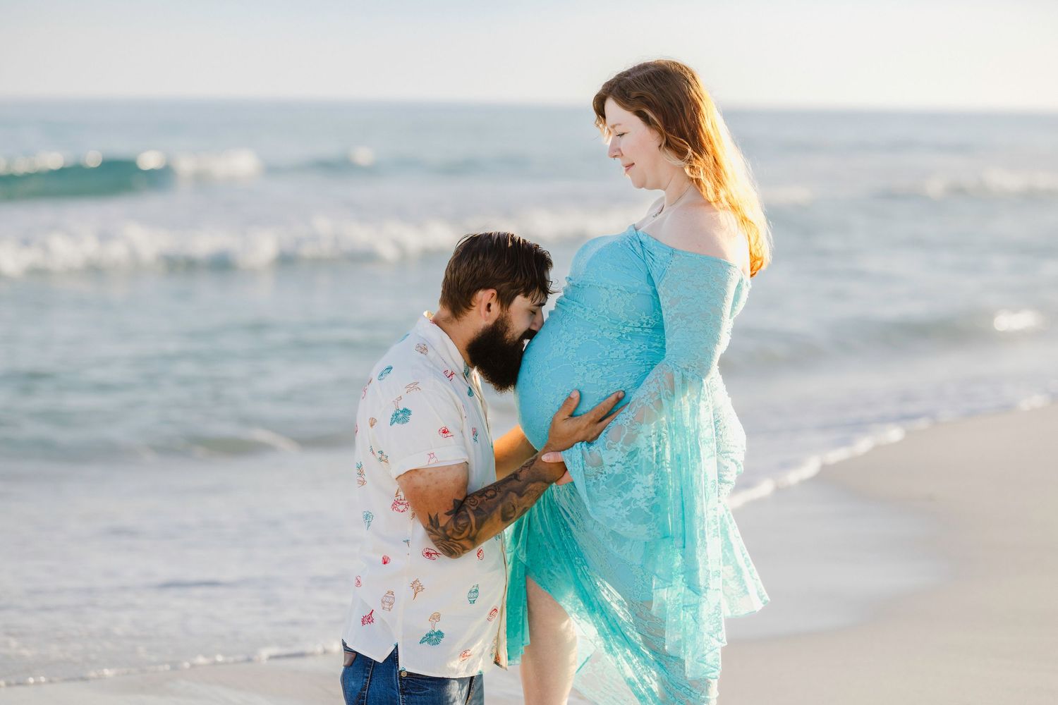 Bearded man tenderly kissing pregnant woman's belly on beach, she wears turquoise gown.