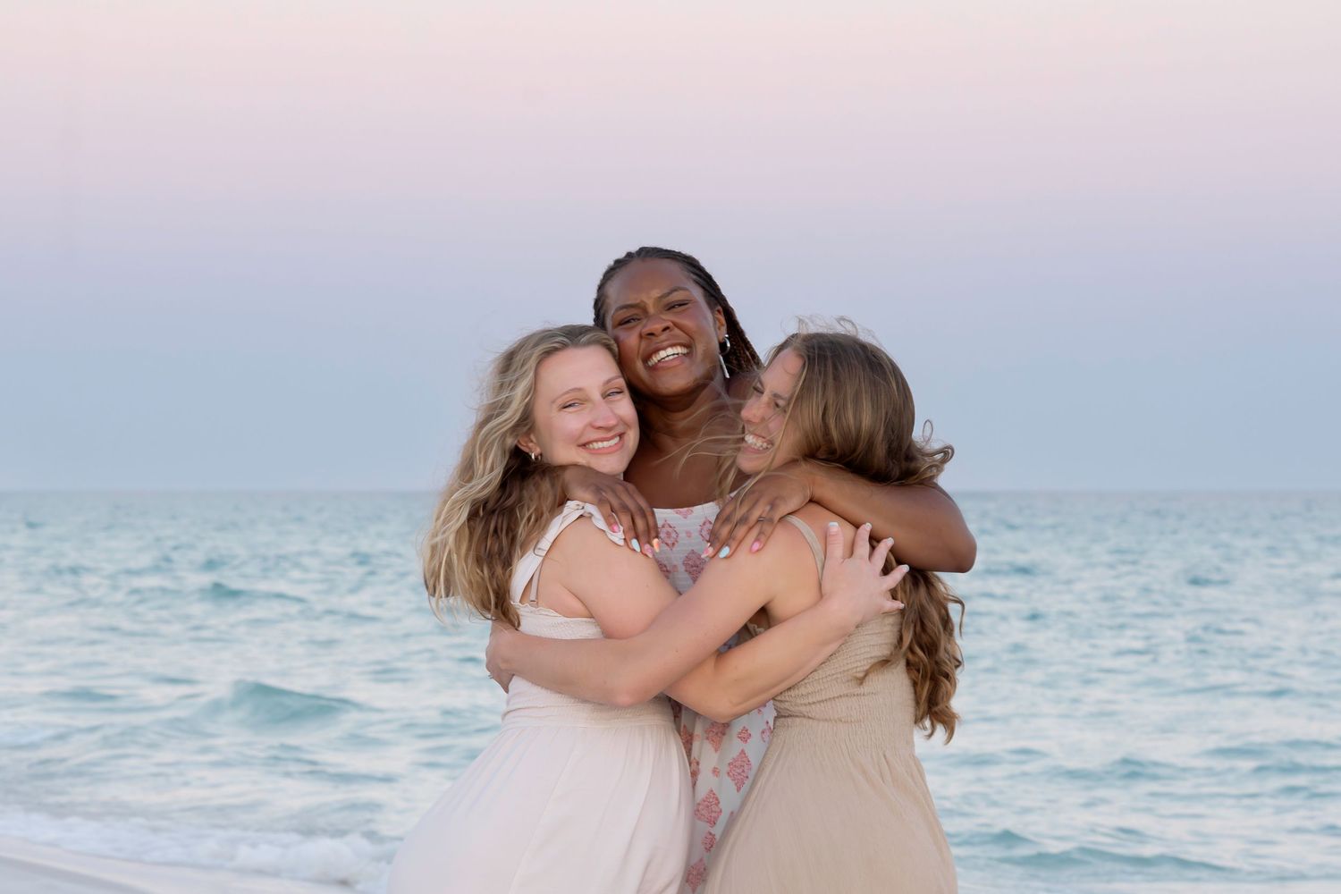 Three women hugging and smiling on a beach at sunset with calm ocean waves in the background.