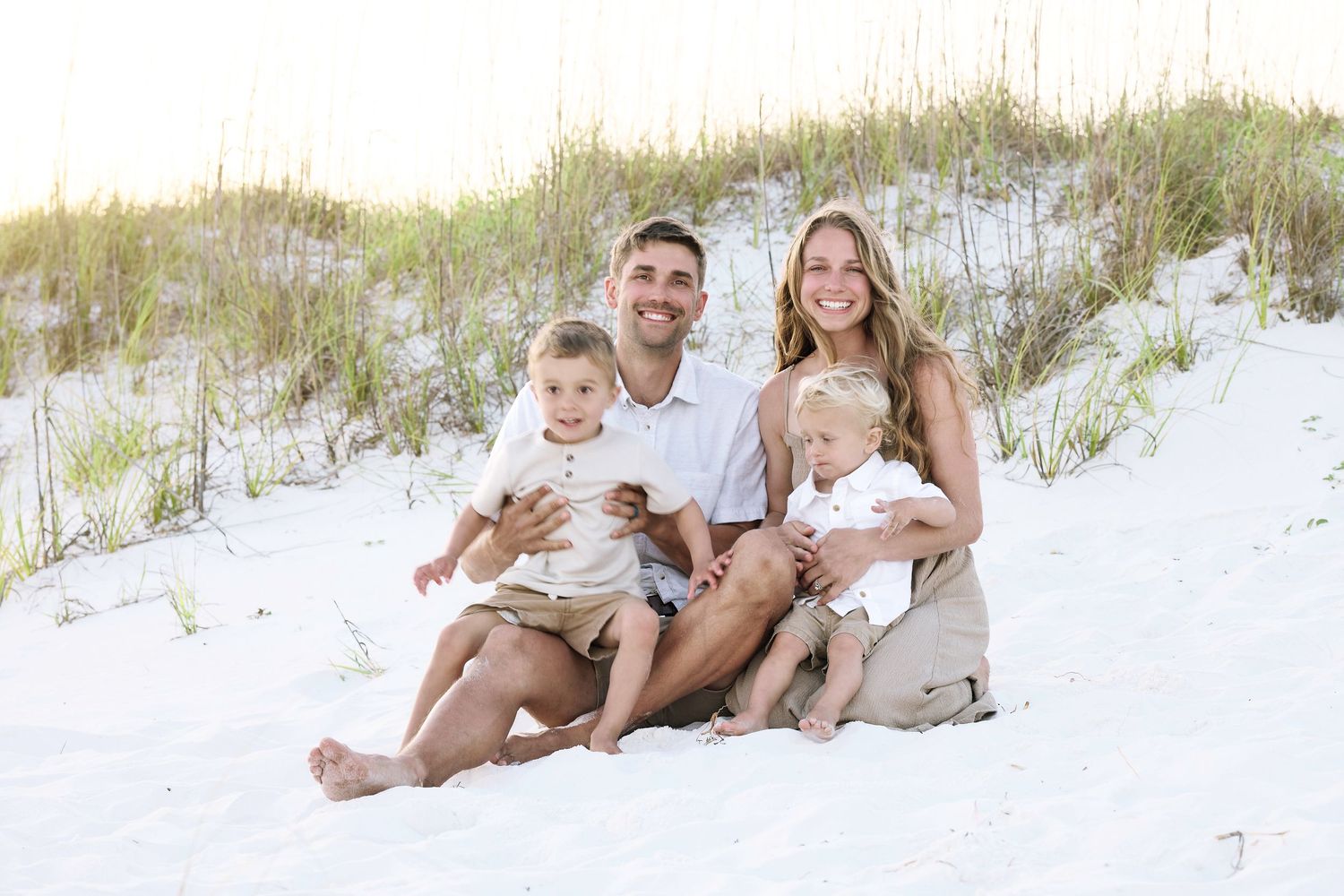 Family of four smiling on white sand beach, parents holding two young children with sand dunes in background.