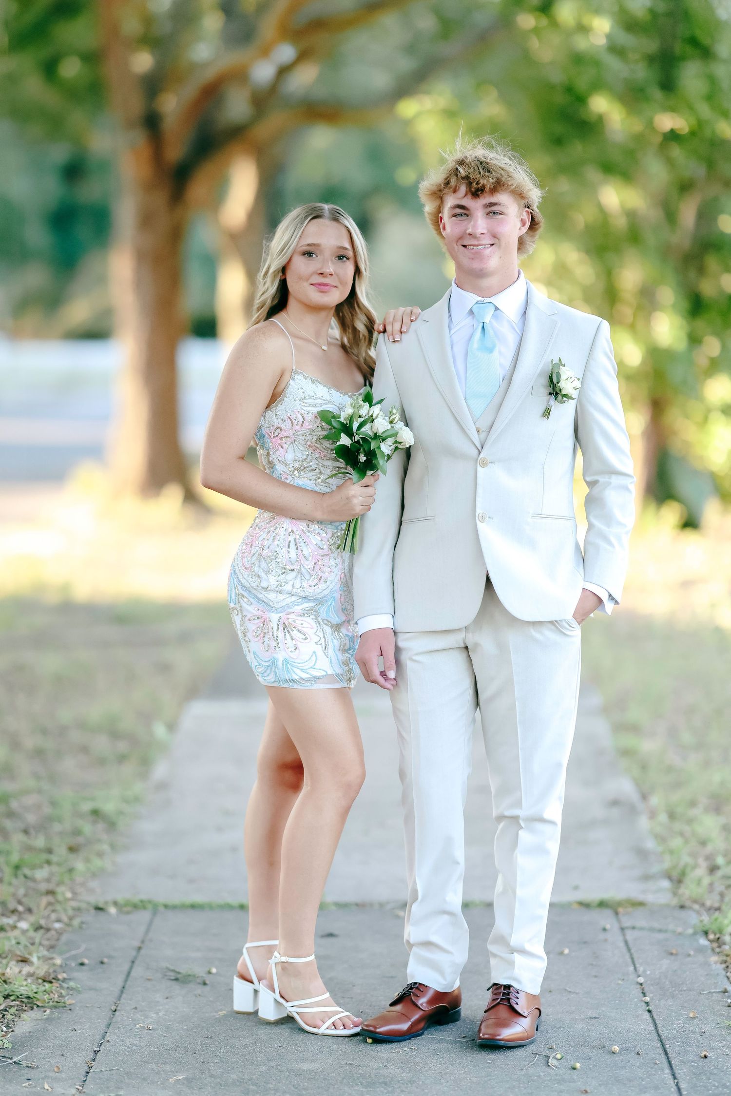 A young couple dressed in prom attire poses outdoors, she in a floral mini dress, he in a cream suit with boutonniere.
