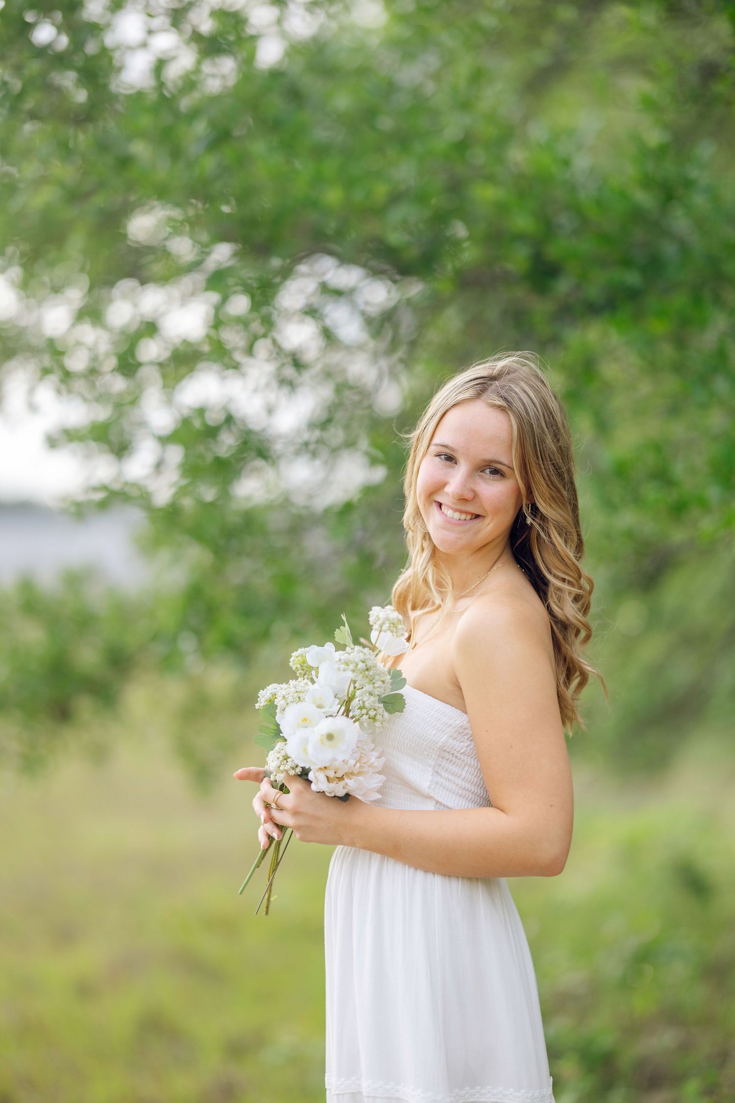 Smiling bride in white strapless dress holding a small white floral bouquet outdoors with green trees in the background.