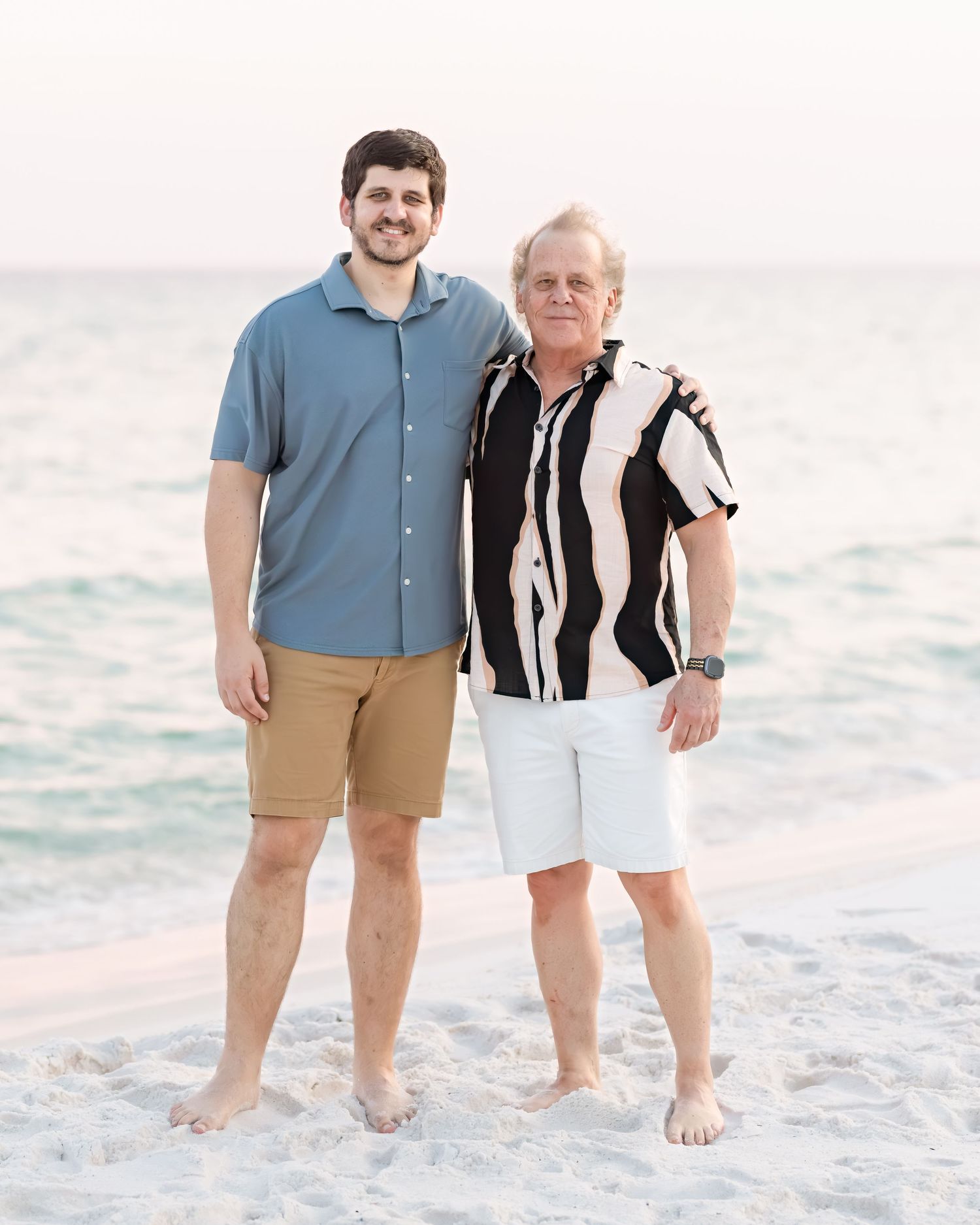 Two men, one younger and one older, pose together smiling on a sunny beach.