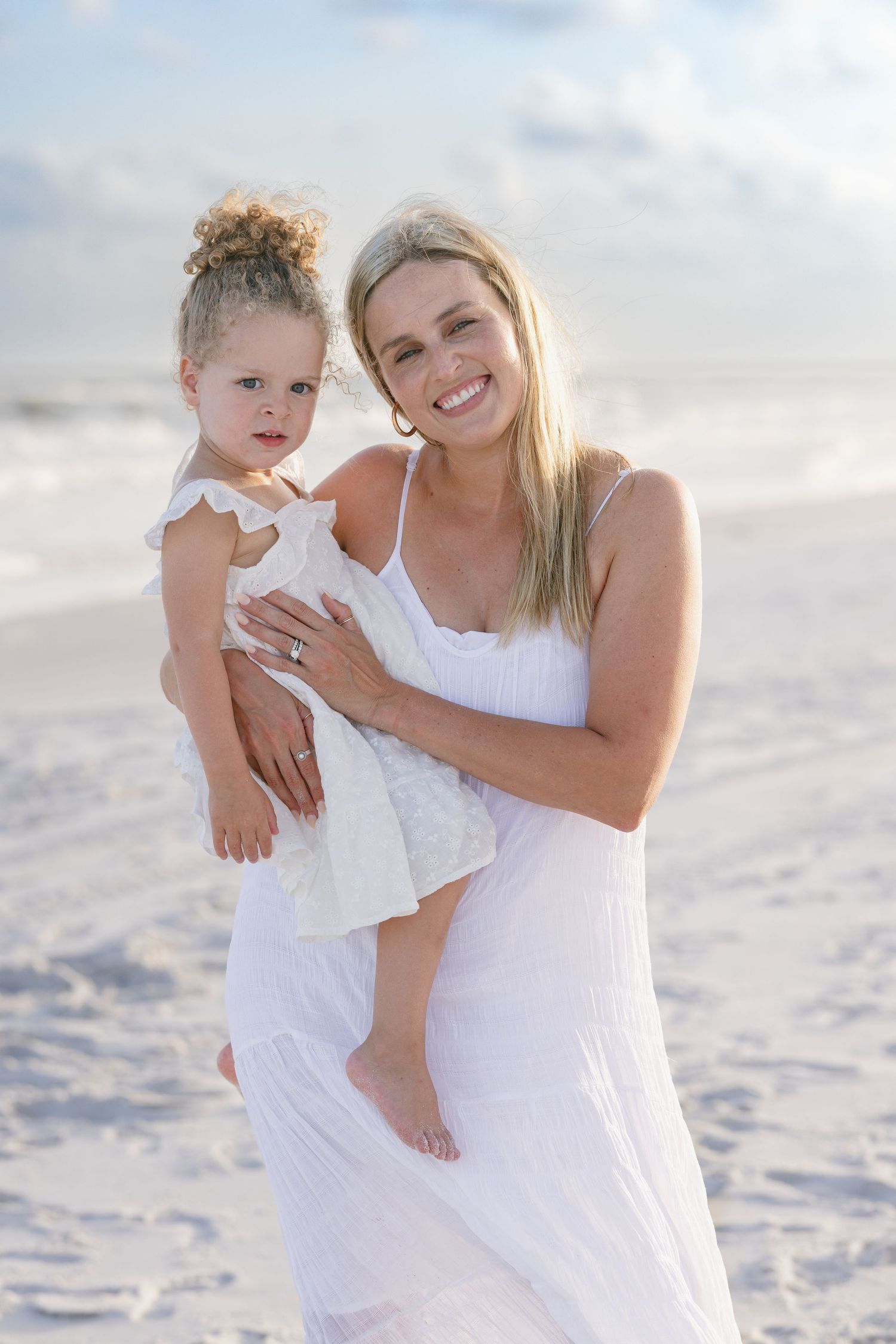 Smiling blonde mother in white dress holding young daughter on sunny beach with ocean waves behind them.