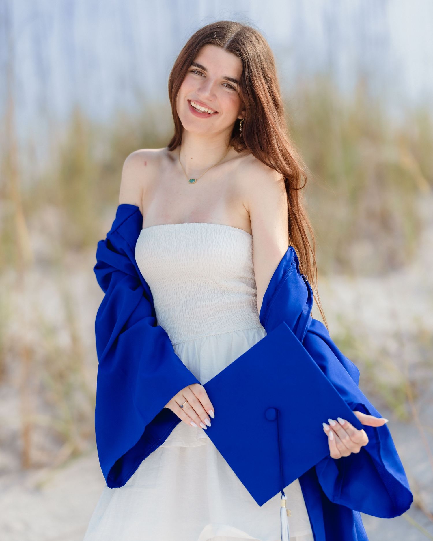 Young woman in white dress holding blue graduation cap and gown, smiling outdoors at a beach setting.