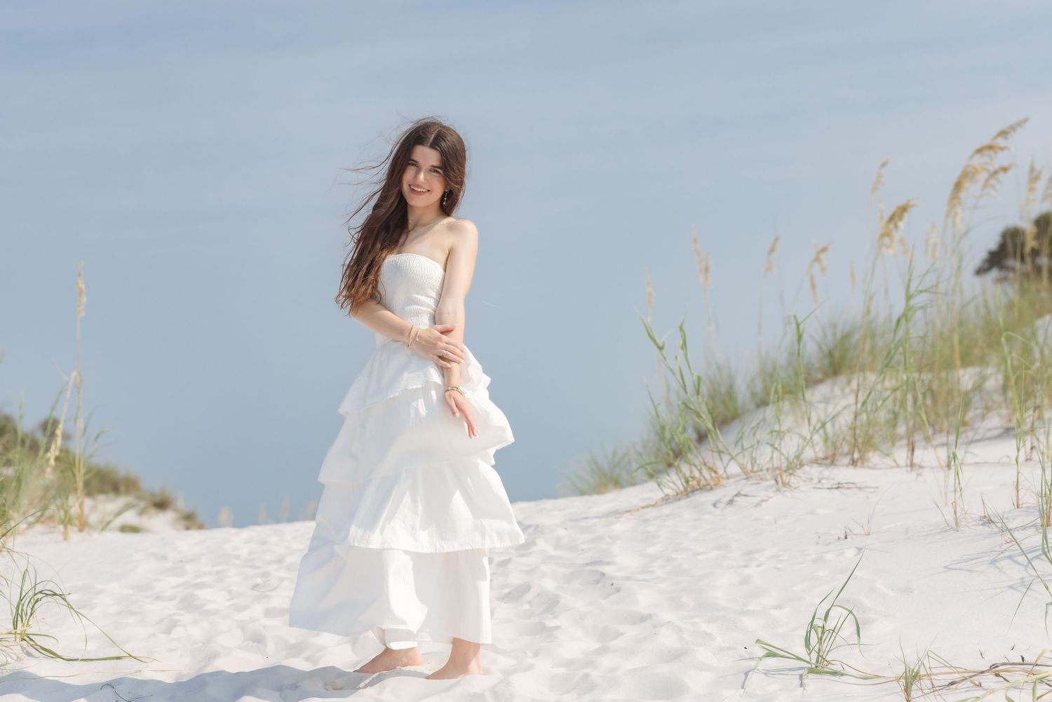 Young woman in white strapless gown poses gracefully on sandy beach dunes with sea oats and blue sky.