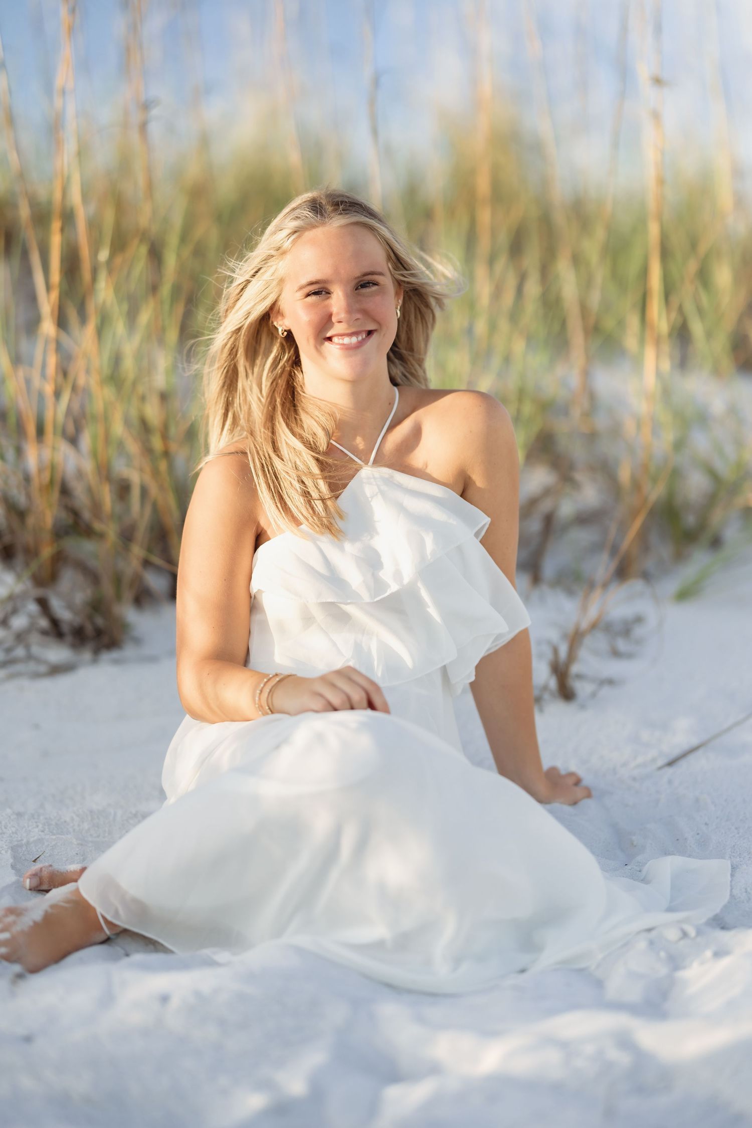 Smiling blonde woman in white ruffled dress sitting on white sand beach with sea oats in background.