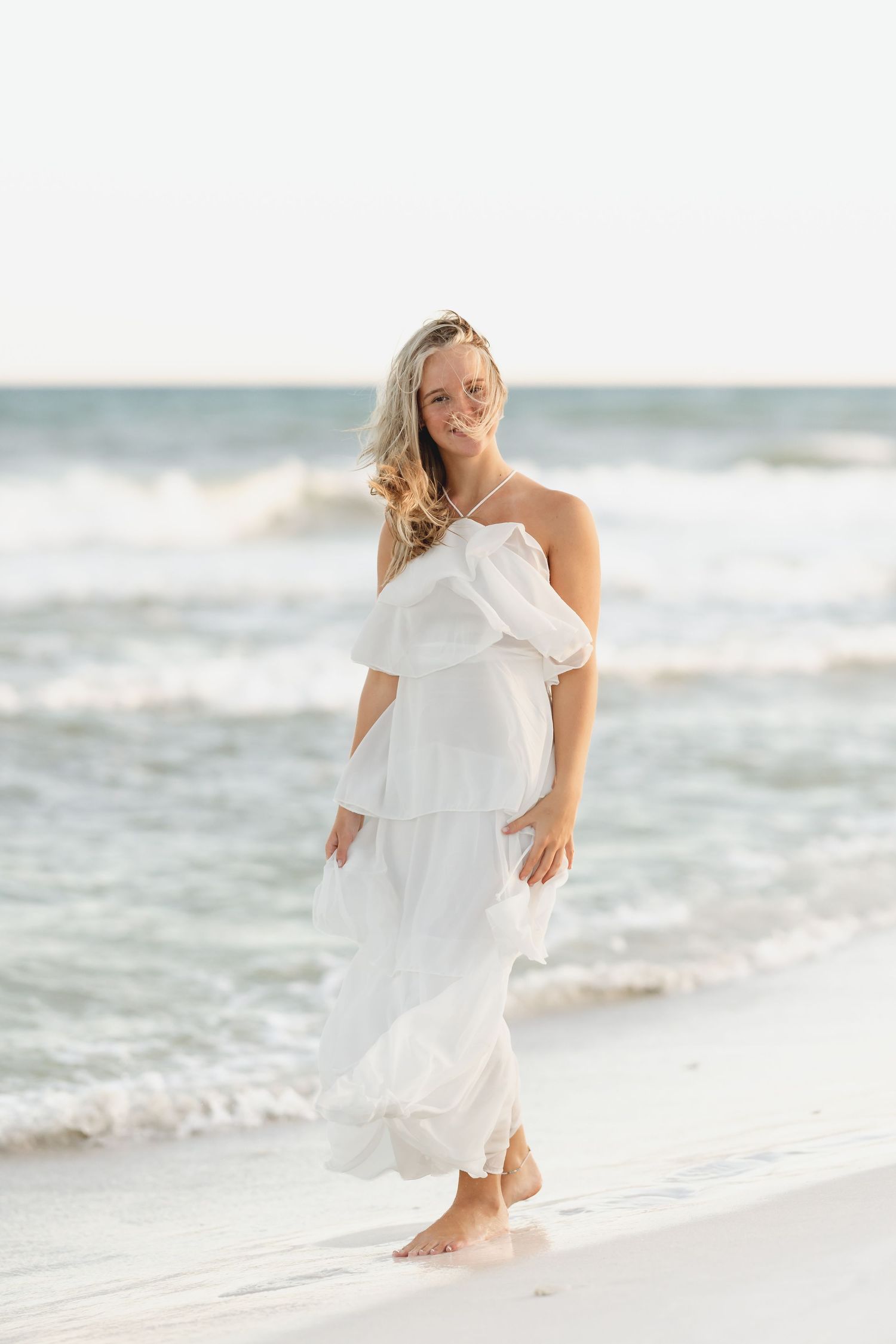 A blonde woman in a flowing white off-shoulder dress walks barefoot on a sandy beach with ocean waves behind her.