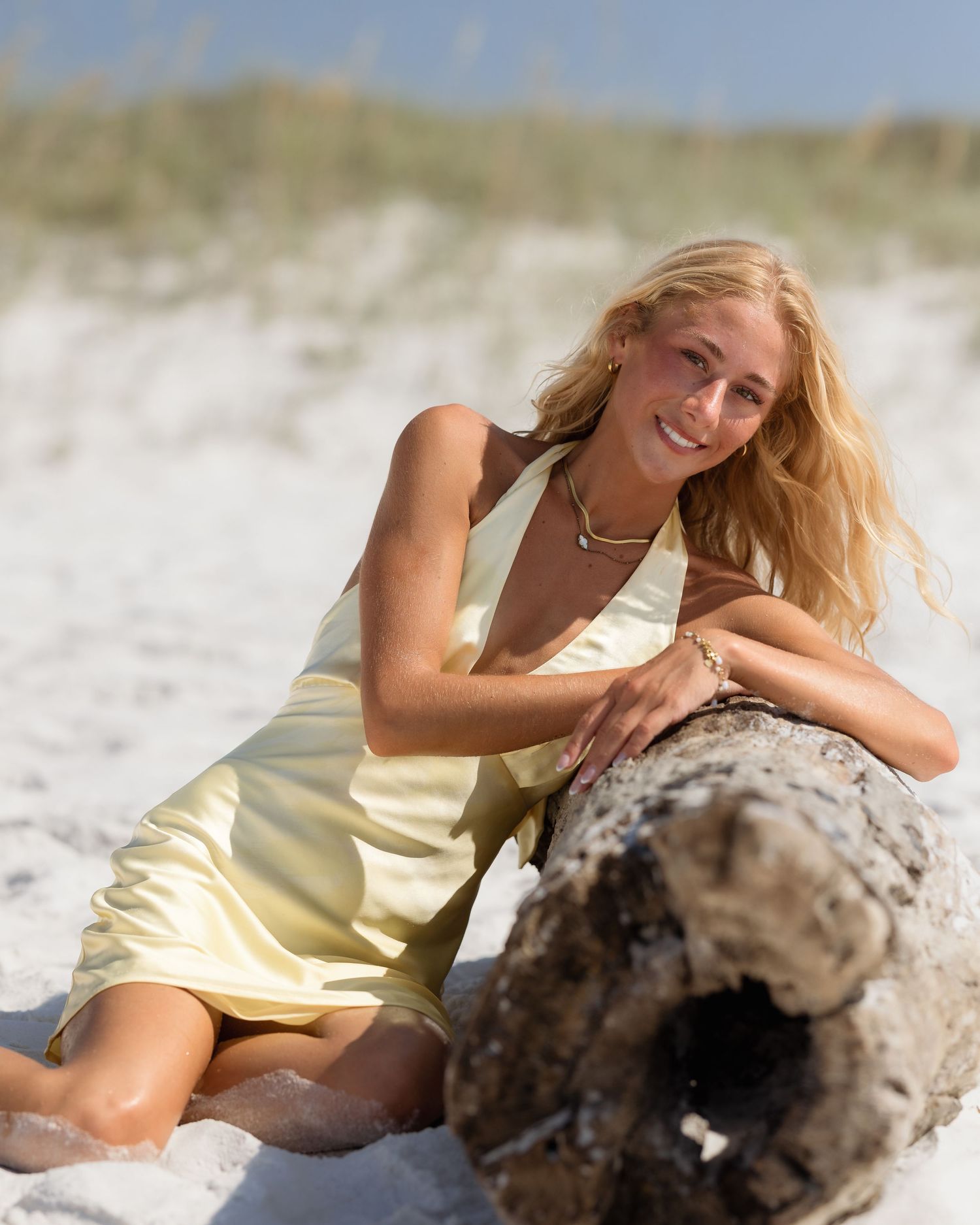 Smiling blonde woman in yellow dress posing with driftwood on a sunny white sand beach.