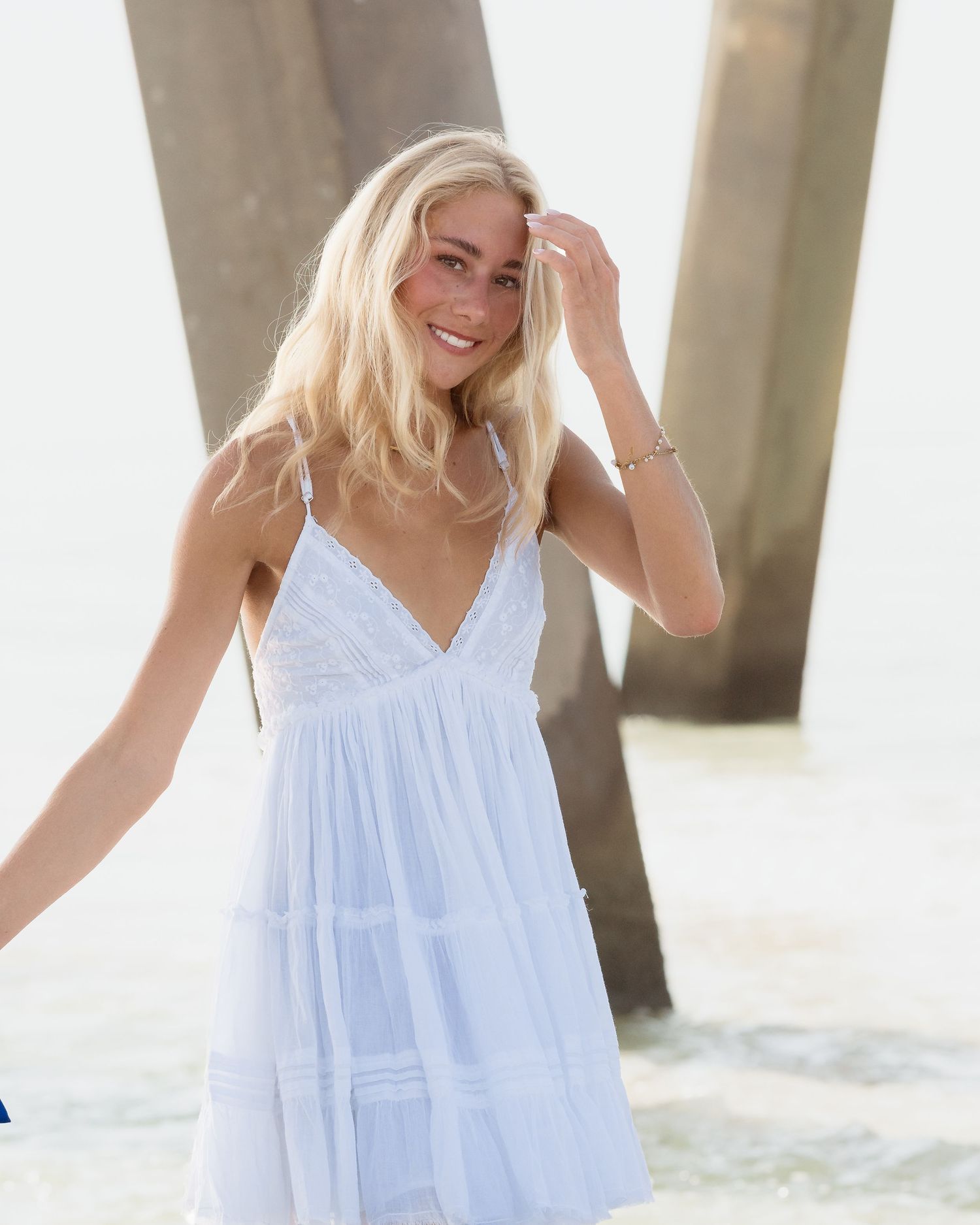 Smiling blonde woman in white v-neck mini dress posing near pier on sunny beach.