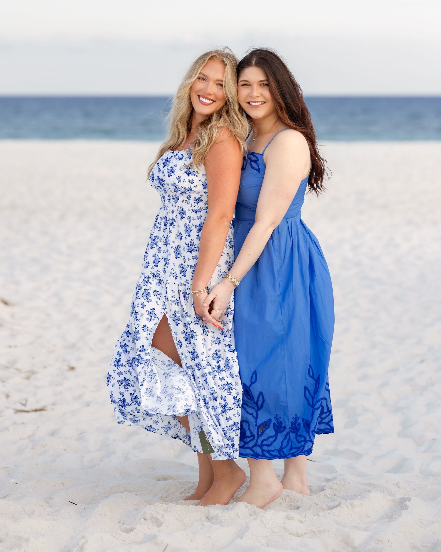 Two women in blue dresses pose together on a sandy beach, smiling and holding hands on a sunny day.