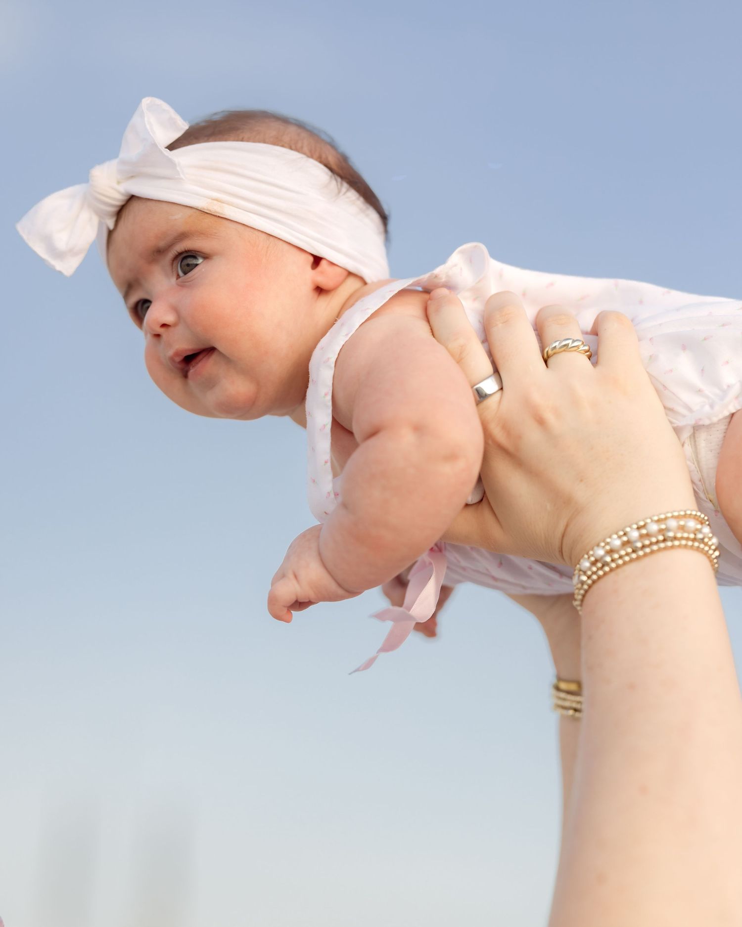 Baby girl in white dress with pink bow being held up by mother wearing gold jewelry on beach.
