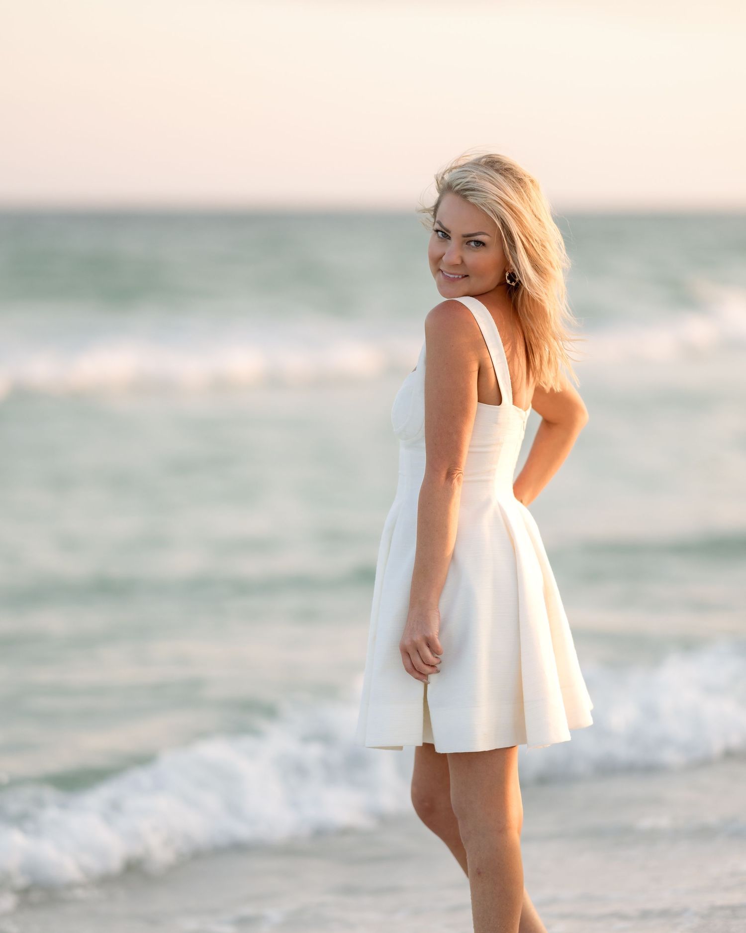 Blonde woman in white dress posing on beach with ocean waves in the background.