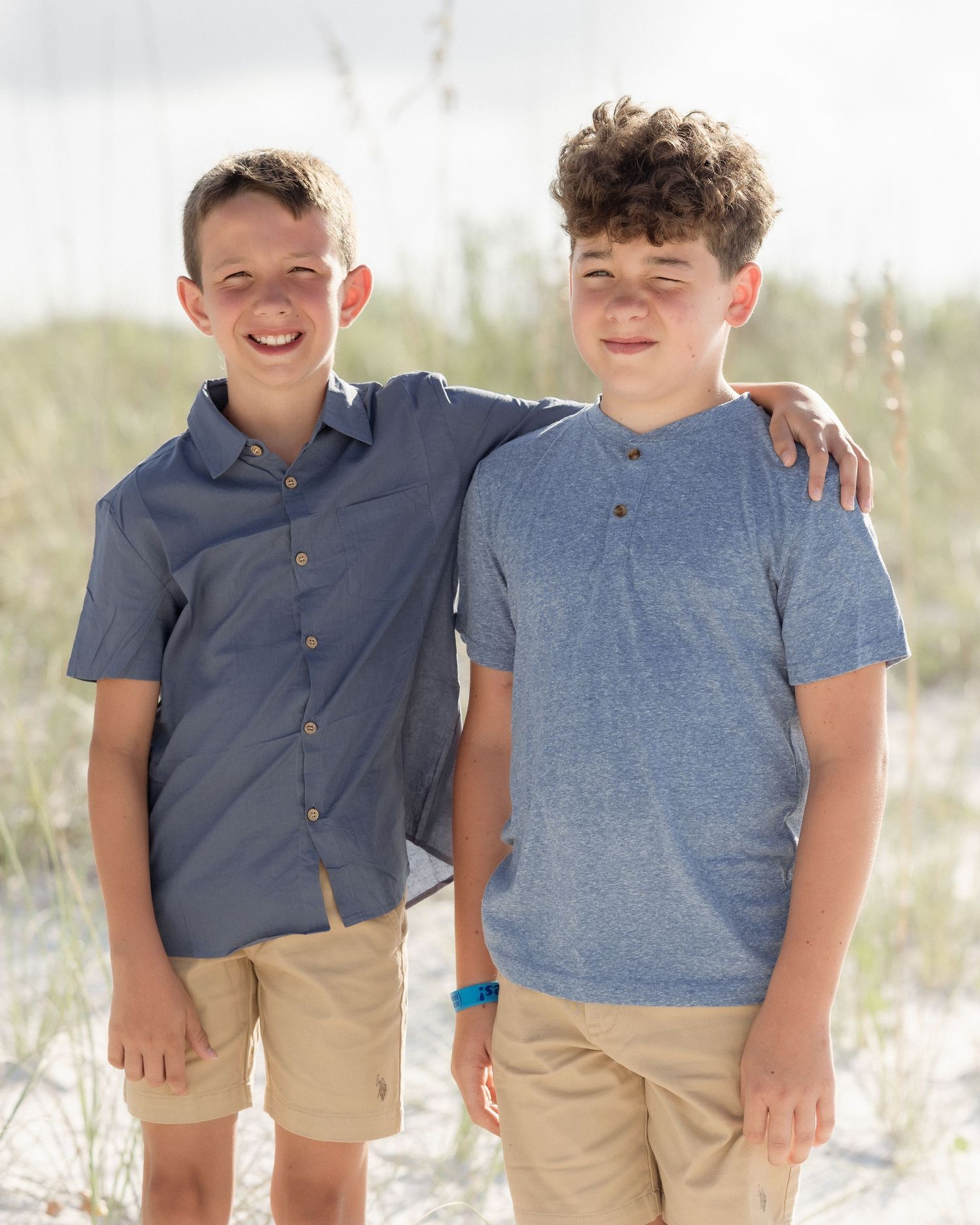 Two smiling teenage boys in blue shirts and khaki shorts pose together on a sandy beach.