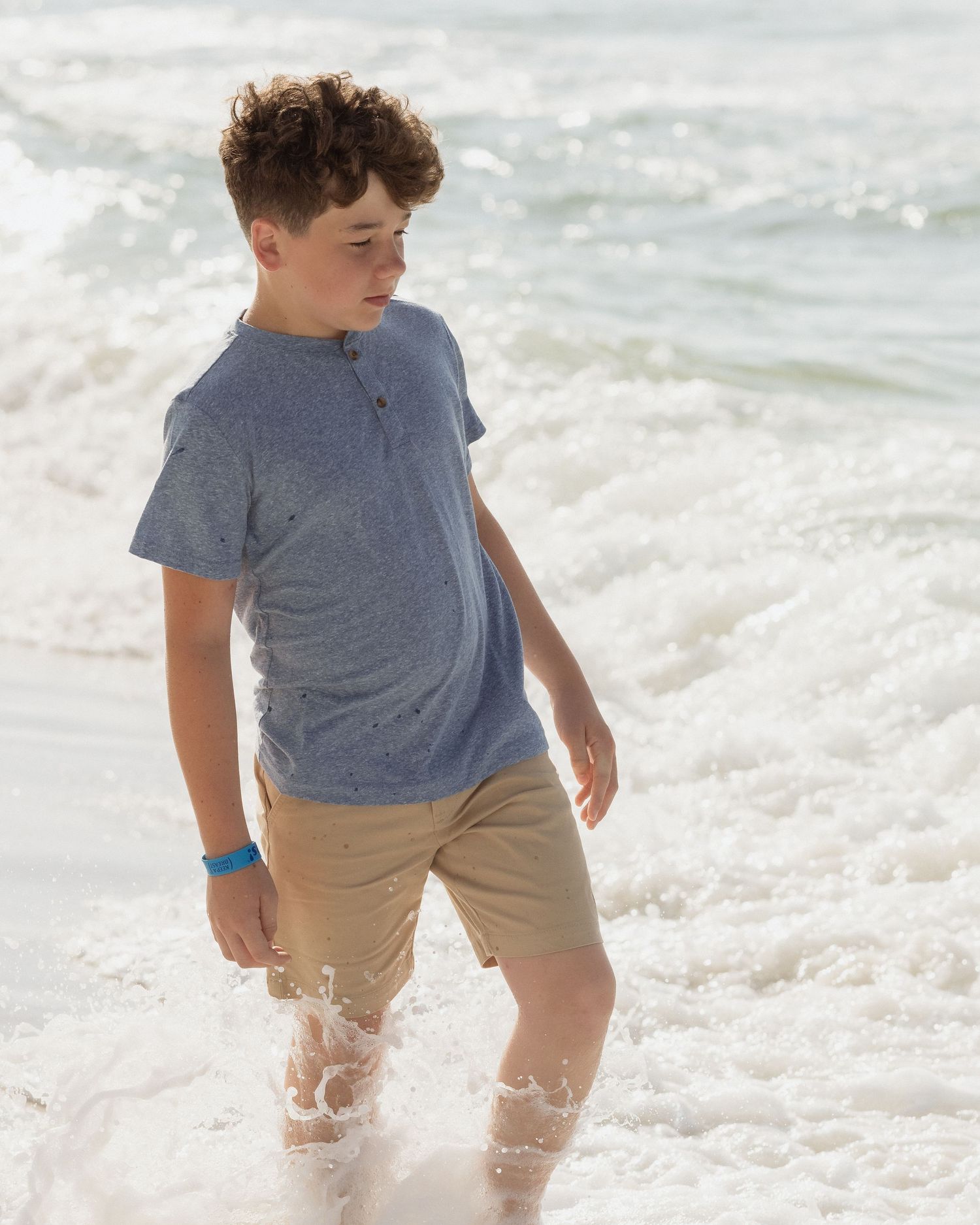 A young boy wades through ocean waves at the beach wearing a blue henley shirt and khaki shorts.