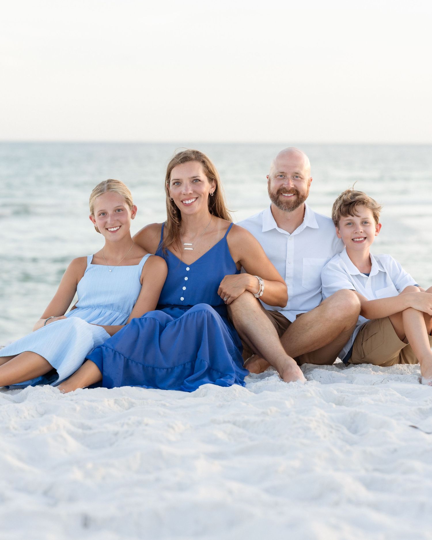 Family of four smiling together on a white sand beach, wearing blue and white outfits with ocean in background.