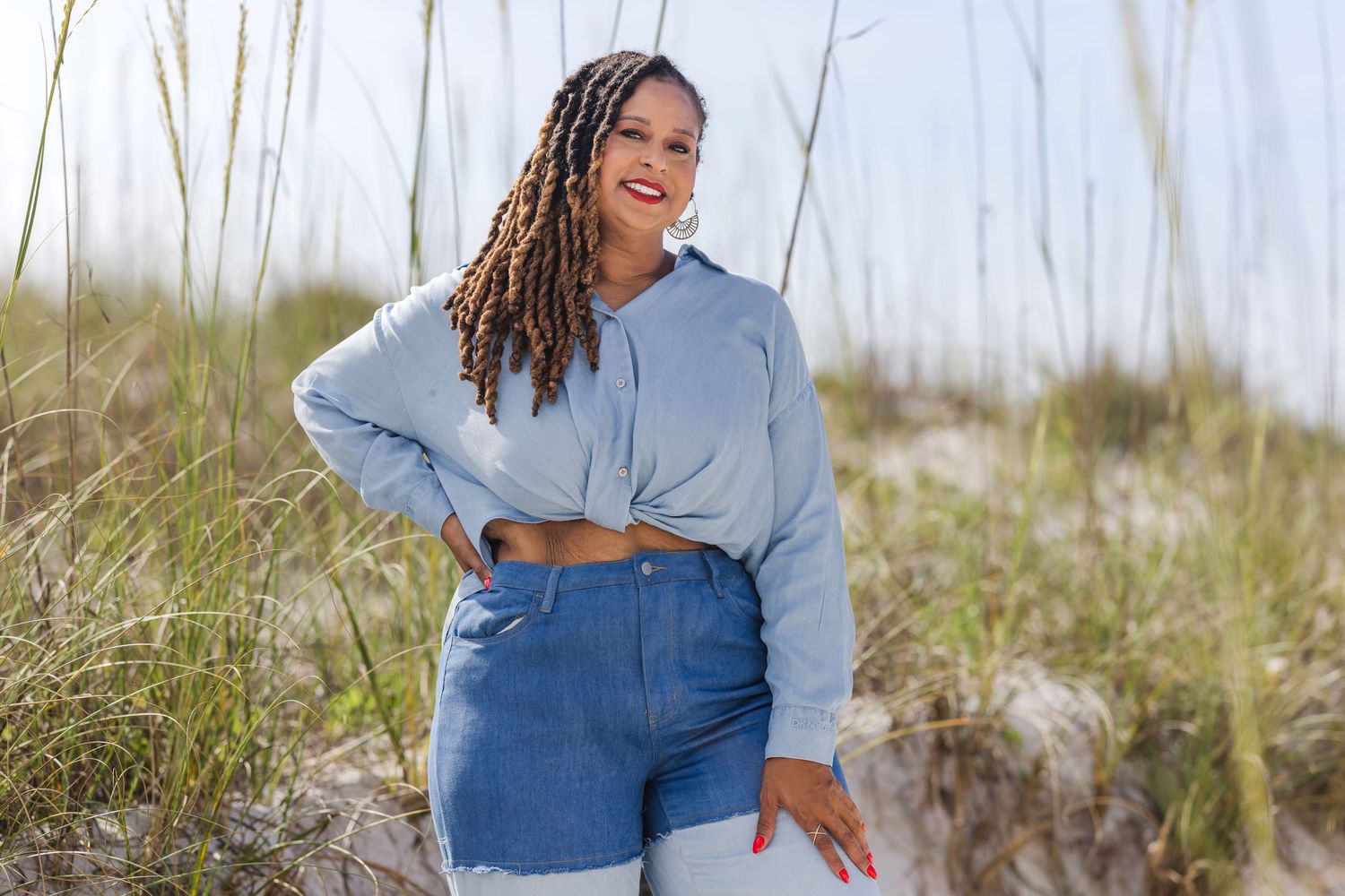 Woman in blue cropped hoodie and jeans smiling on sandy beach dunes with sea oats in background.