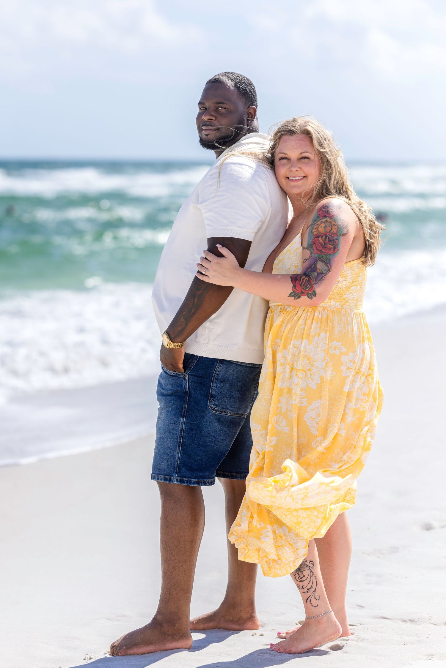Interracial couple embracing on a sunny beach, woman in yellow floral dress with tattoos, man in white shirt and denim shorts.