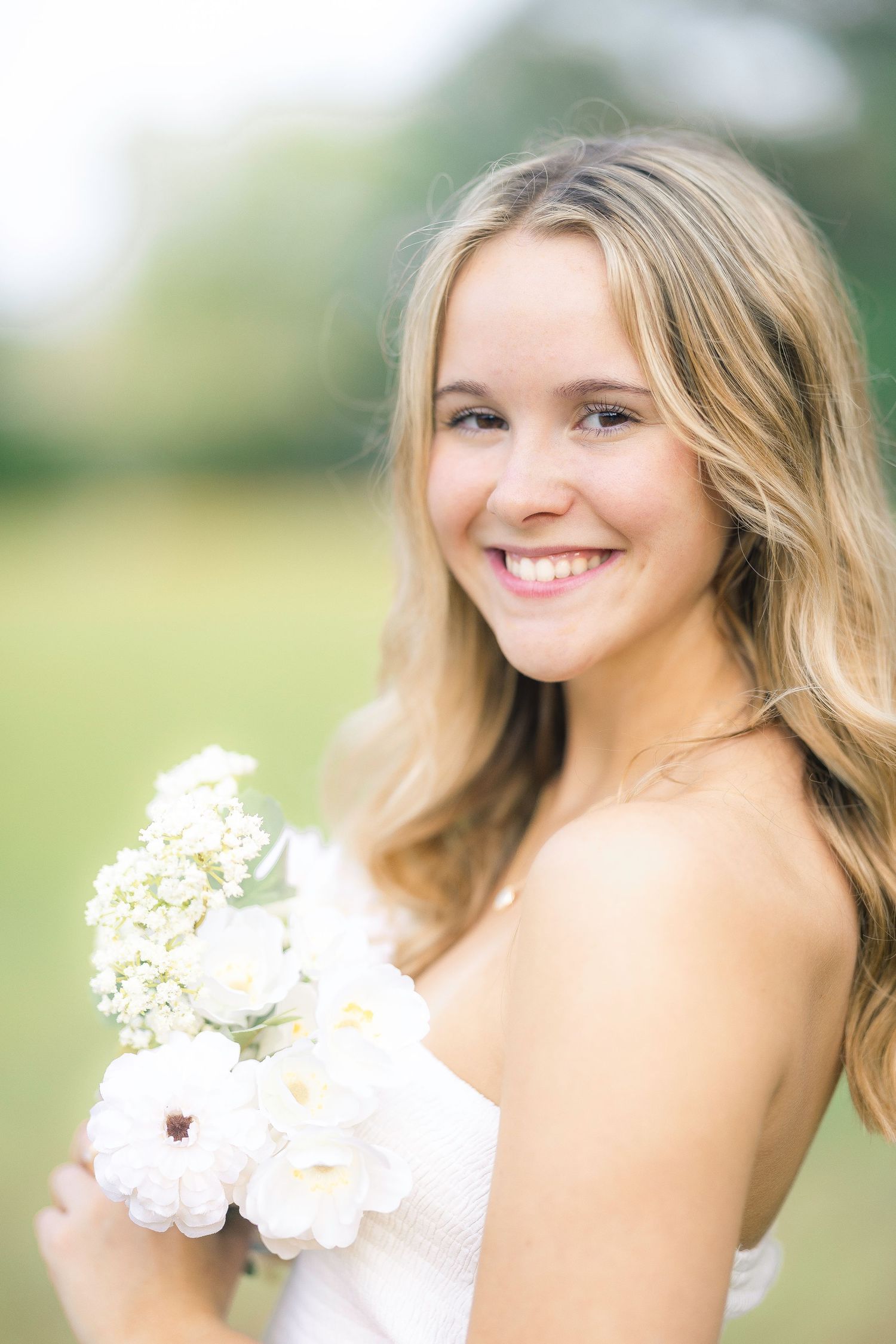 Smiling young woman with blonde hair holding white flowers outdoors in a soft, natural light setting.