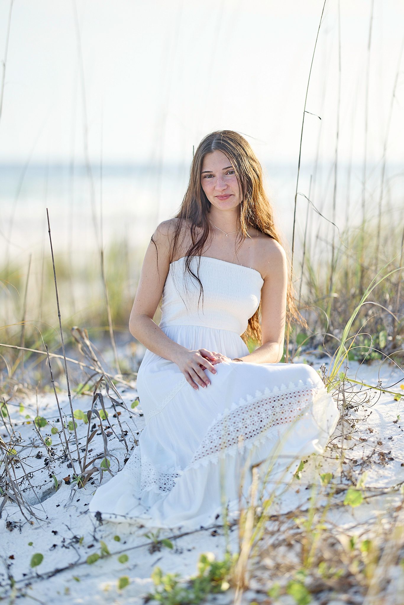 Young woman in white sundress sitting on sandy beach dunes with sea grass, bathed in warm sunlight.