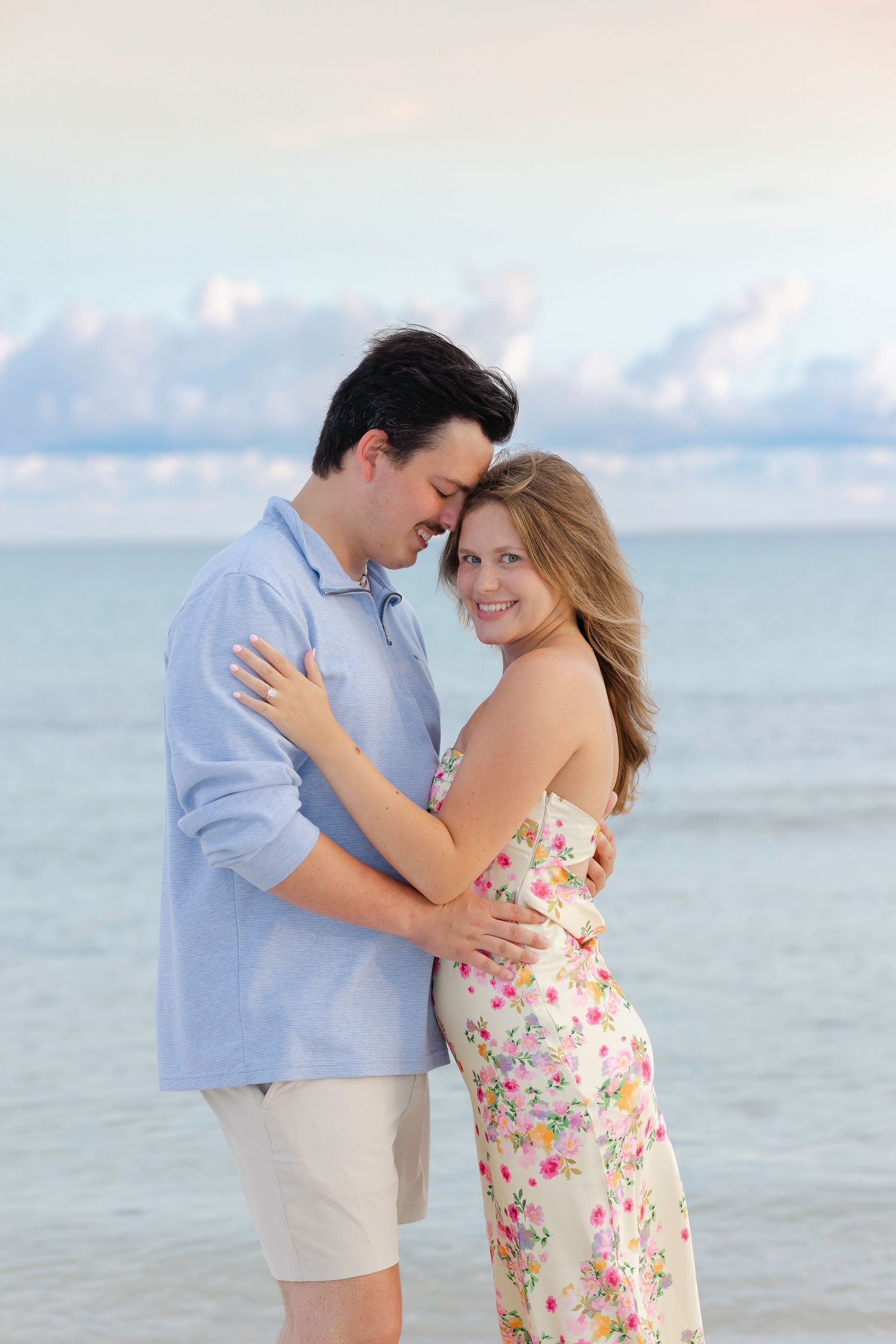 A couple embraces romantically on a beach at sunset, woman in floral dress smiling at camera, man in blue shirt.