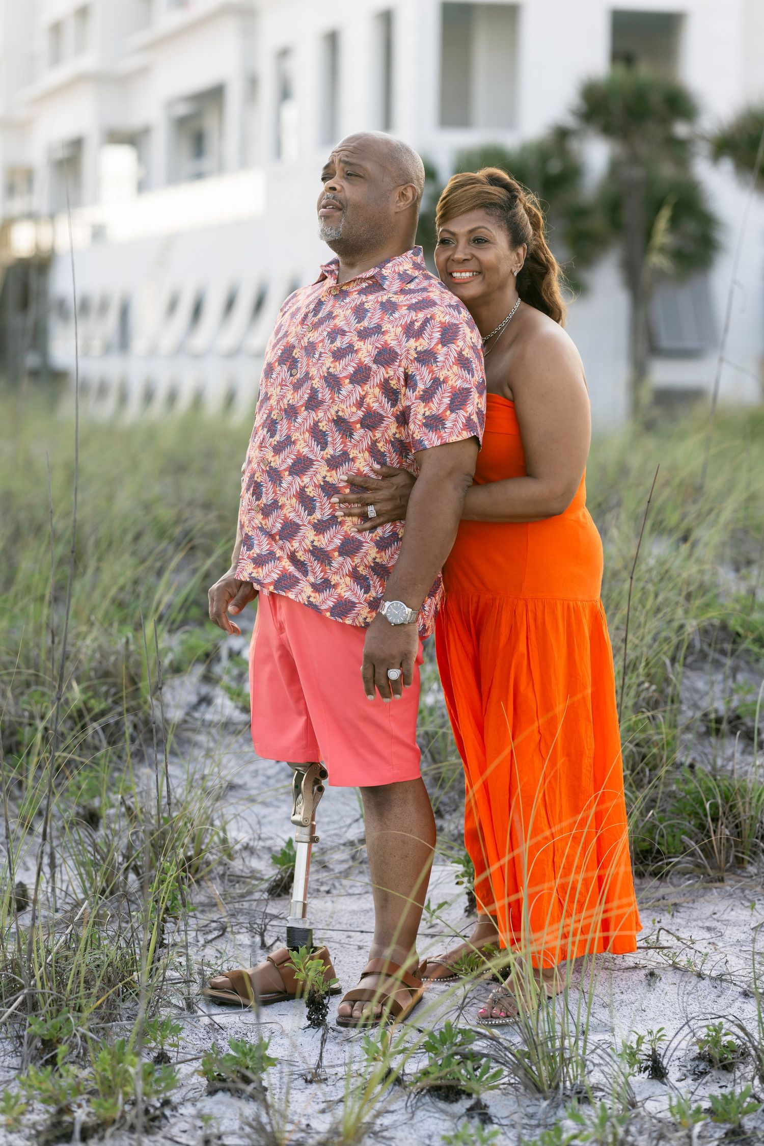 Couple posing on sandy beach path, woman in orange maxi dress embracing man in pink floral shirt with prosthetic leg.