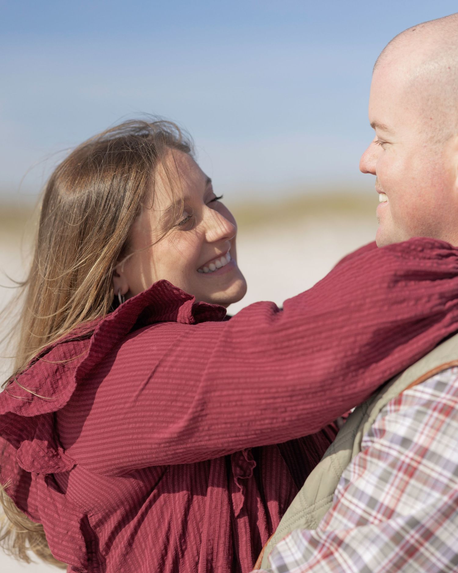 A smiling couple embraces on a sunny beach, the woman in a burgundy top gazing lovingly at the man.