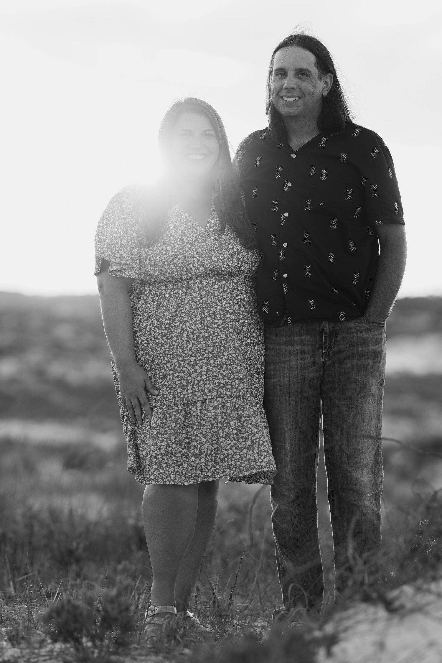 Black and white photo of a smiling couple standing together outdoors at sunset in a field.