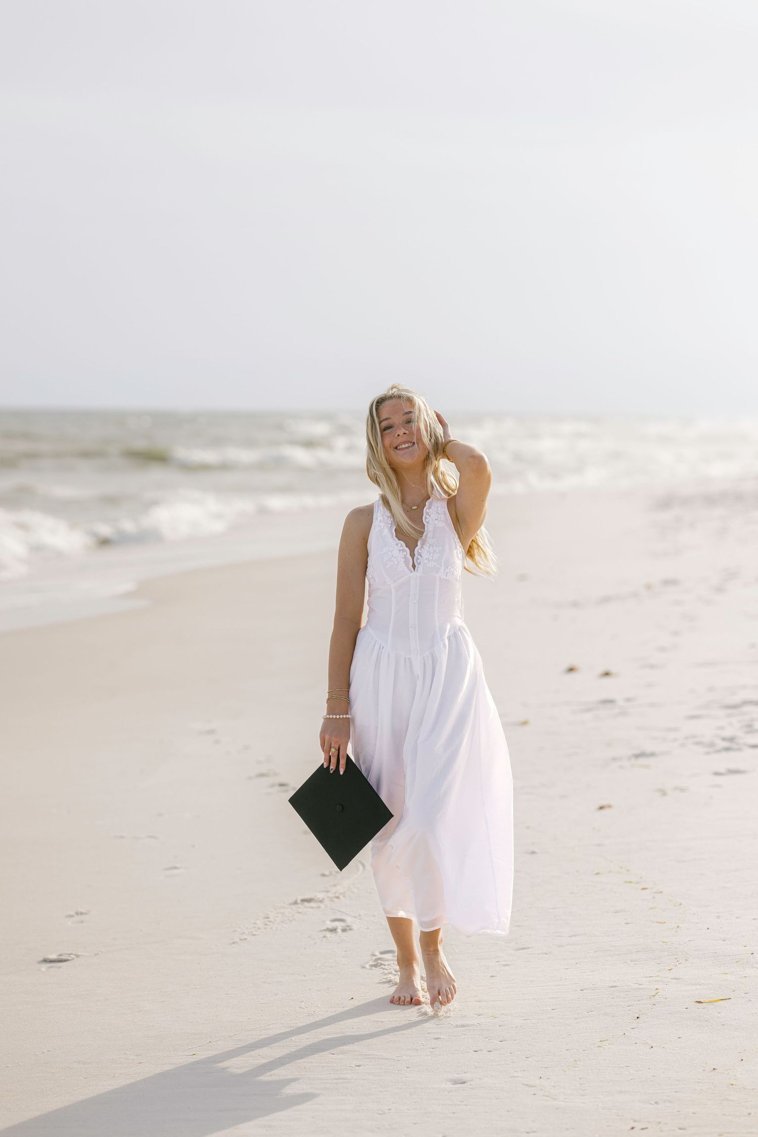 Smiling blonde woman in white maxi dress holding graduation cap walks barefoot on sunny beach.