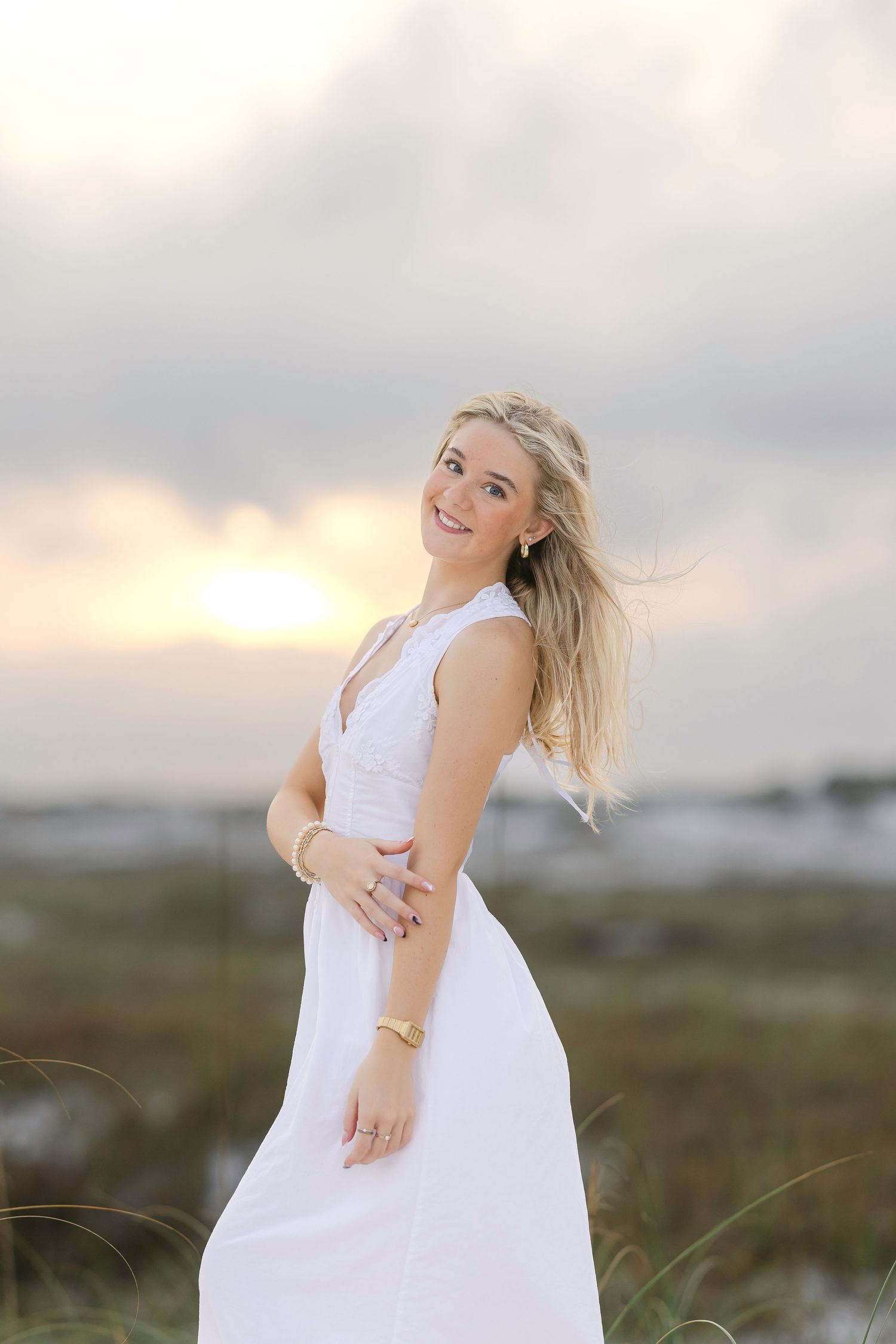 Blonde woman in white maxi dress smiles at sunset on beach with wind-blown hair and gold jewelry.